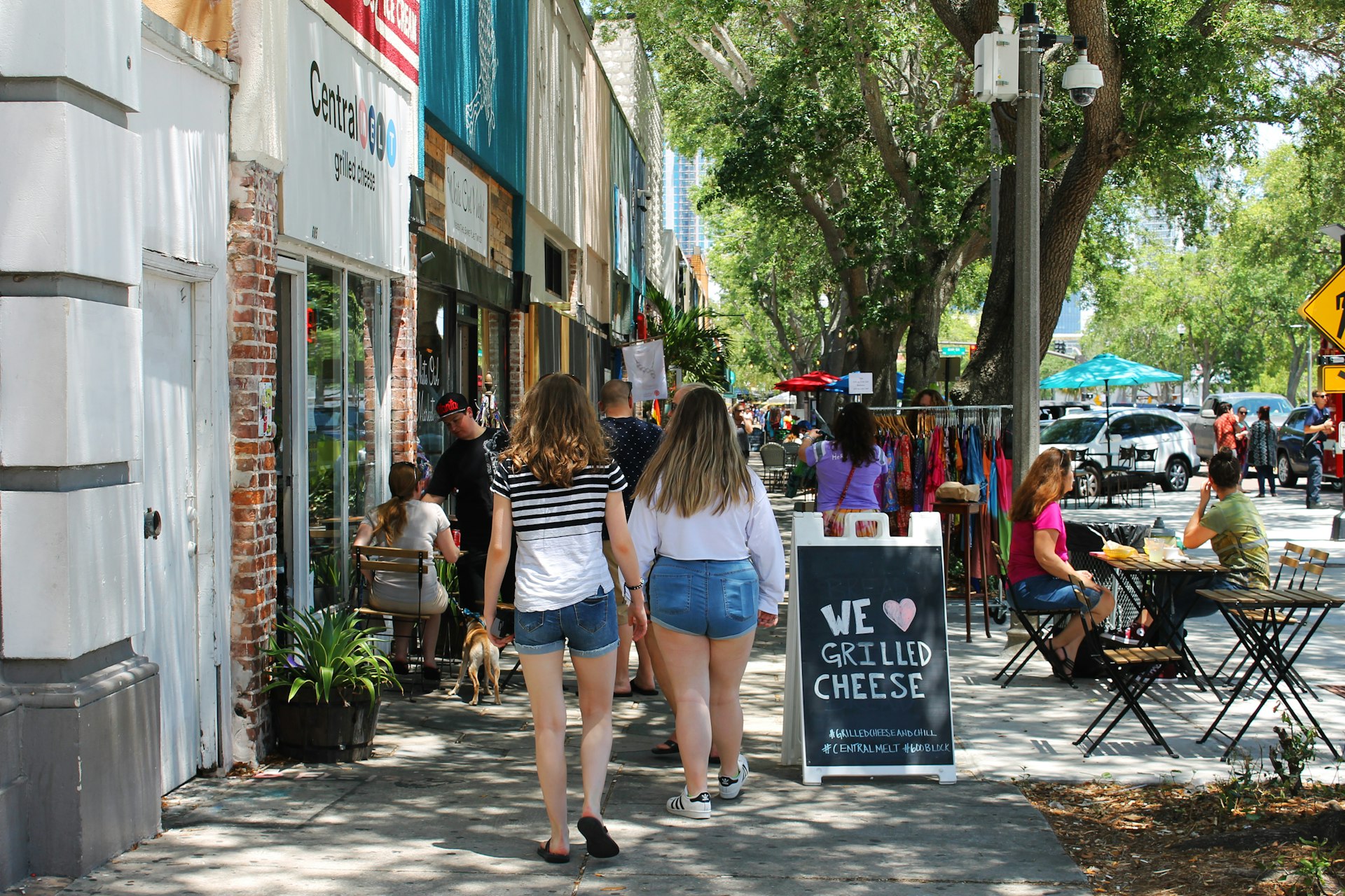 Two young women with dirty blonde hair in blue jean shorts and white tops walk past rows of shops in St. Pete. where a couple are sitting at cafe tables outside a restaurant advertising grilled cheese on a sandwich board sign. Racks of colorful clothes sit on the sidewalk in the background, where another woman is browsing.