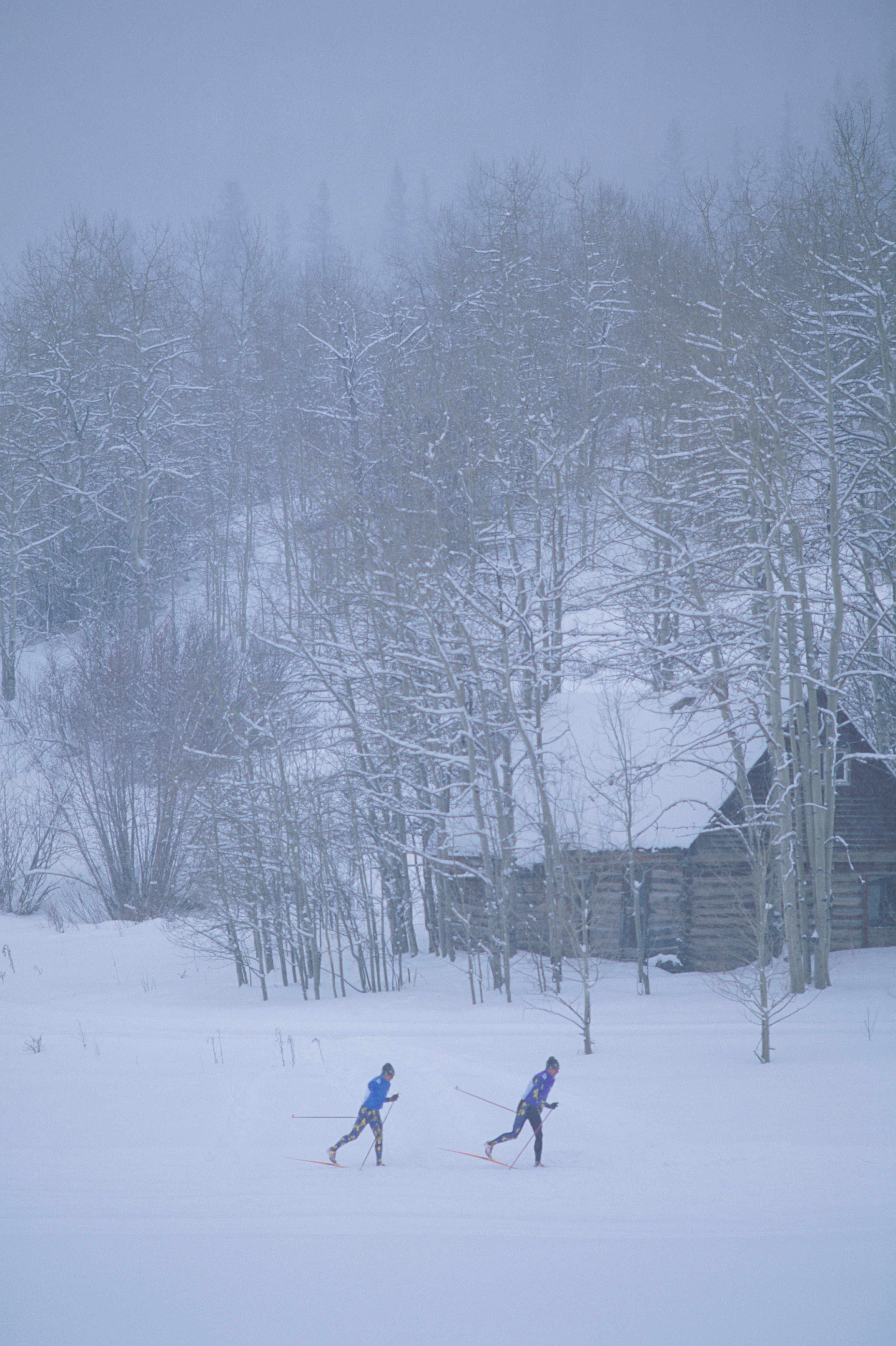 Two men nordic ski across a snowy meadow in front of a log cabin in the snow