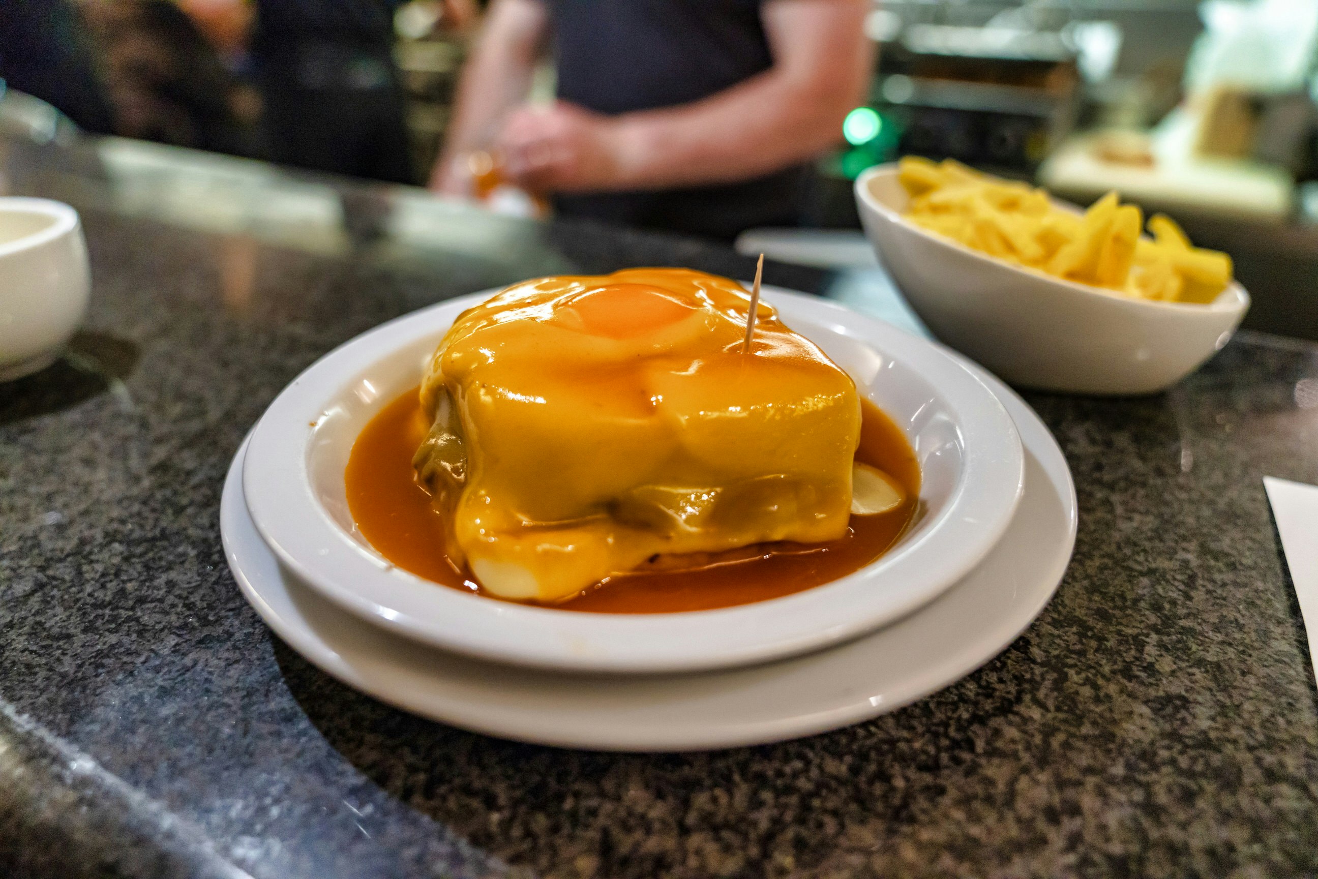 A francesinha in on a white plate, which sits on top of a bar. It's smothered with sauce and behind it sits a bowl of fries. A member of staff is out of focus, behind the bar.