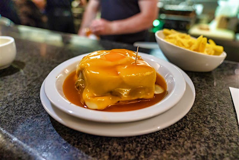 A francesinha in on a white plate, which sits on top of a bar. It's smothered with sauce and behind it sits a bowl of fries. A member of staff is out of focus, behind the bar.