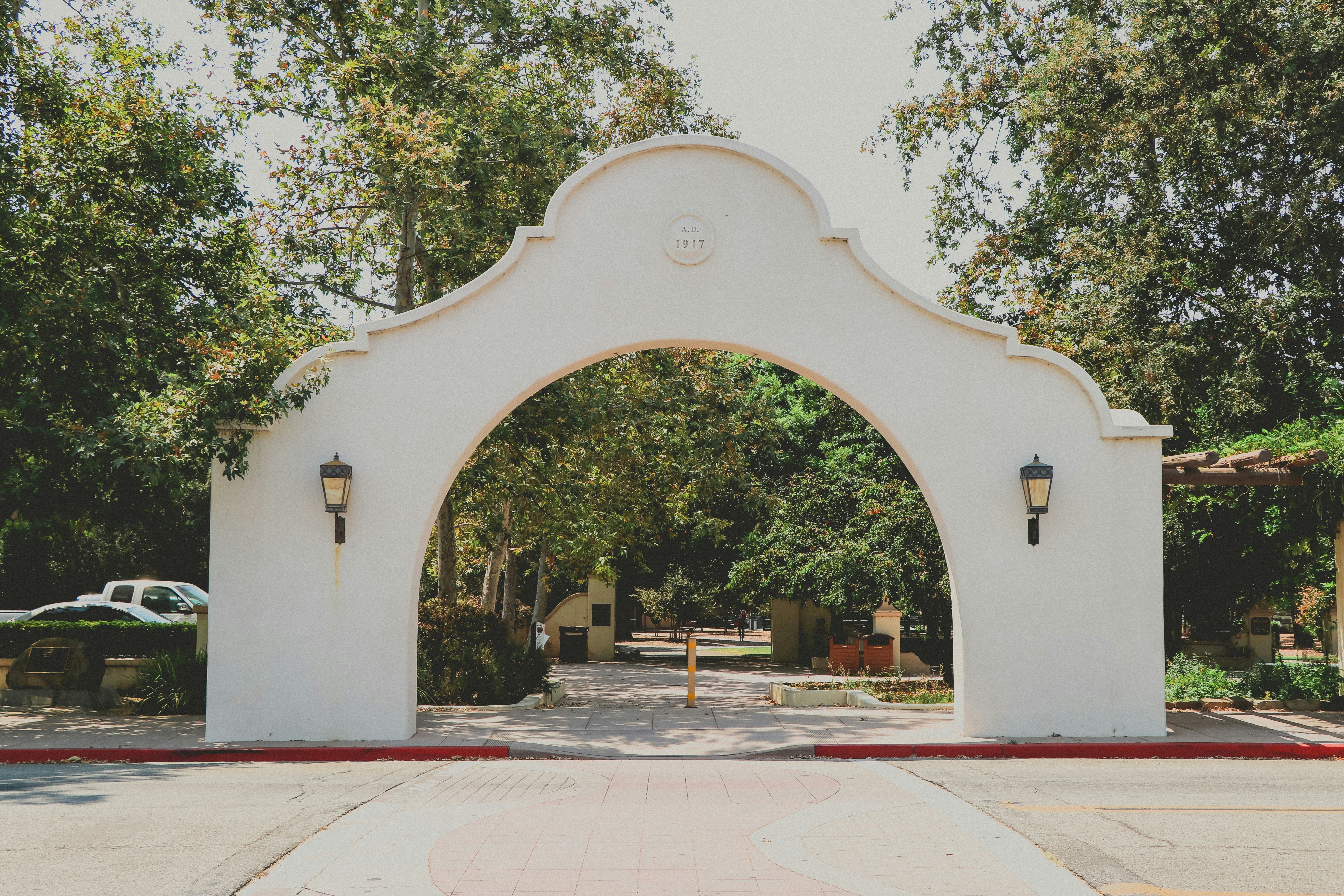 A white mission-style adobe arch with a scalloped top and two black iron lantern sconces marks the entrance to Libbey Park in Ojai, California, full of large green trees and a paved walkway from the viewer straight back in the frame.
