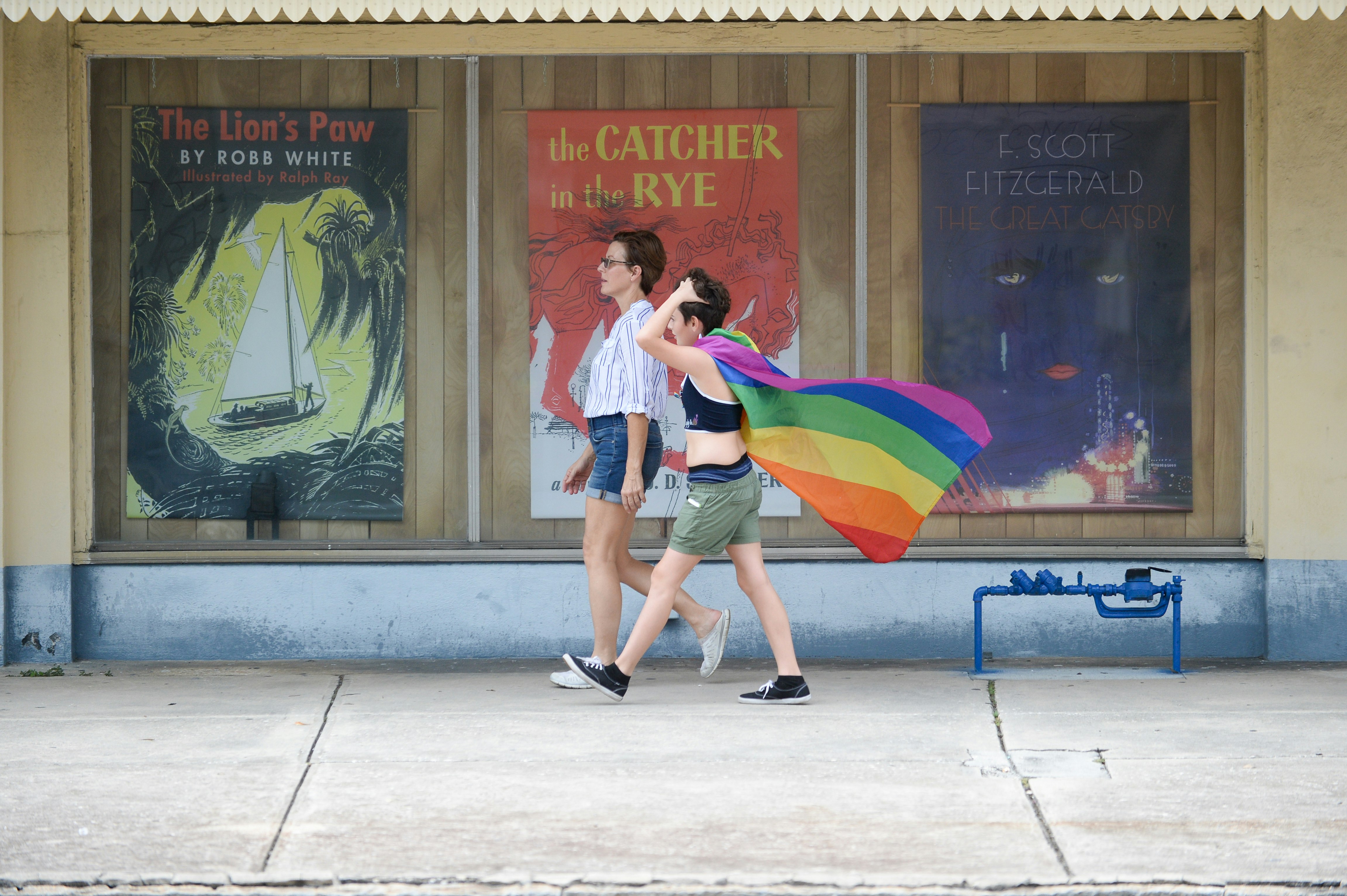 Two short-haired brunette women, one in jean shorts and a white button-down and the other in olive shorts and a black crop top, wearing a Pride flag as a cape, walk past the enlarged covers of classic books, including Catcher in the Rye, that decorate the exterior of Haslam's Bookstore