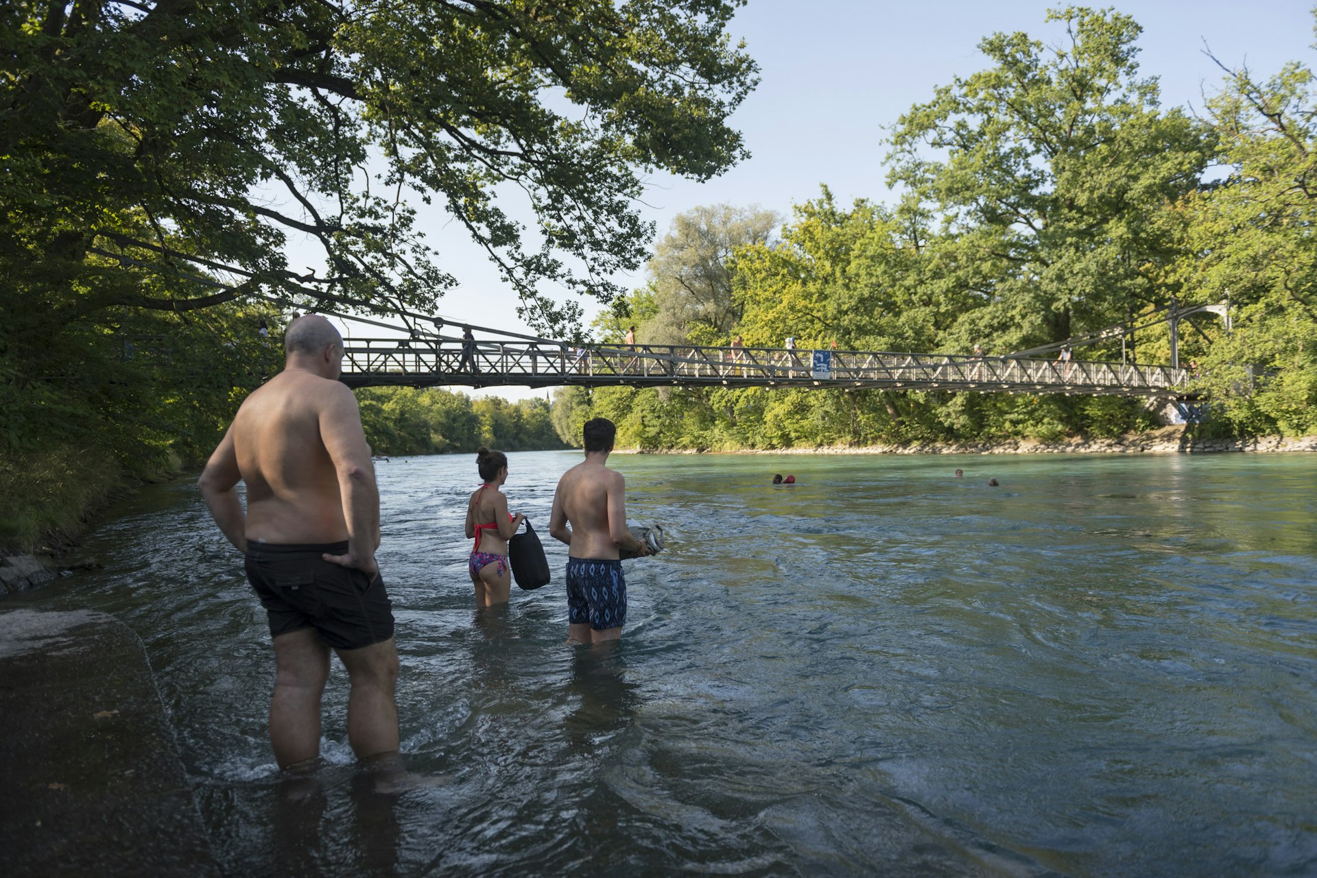 A group of people stand on the bank of a river and look towards a pedestrian bridge, underneath which people are swimming.