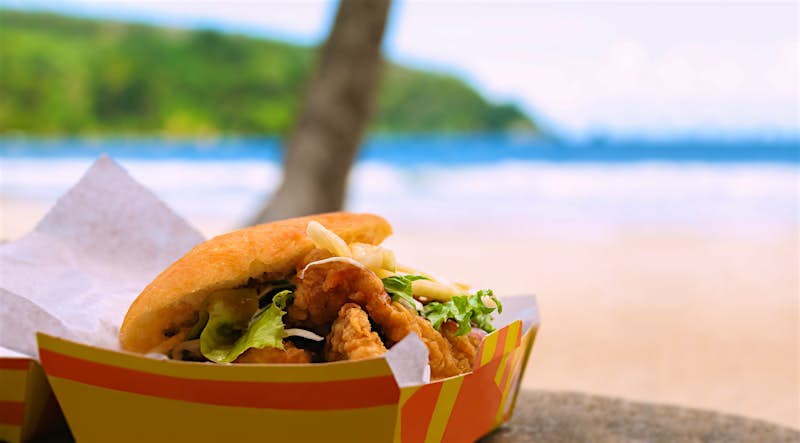 An open cardboard carton containing a flatbread stuffed with pieces of deep-fried shark lettuce and other fillings, is sitting on a table outside. The trunk of a plam tree and the beach and sea beyond, is out of focus in the background 