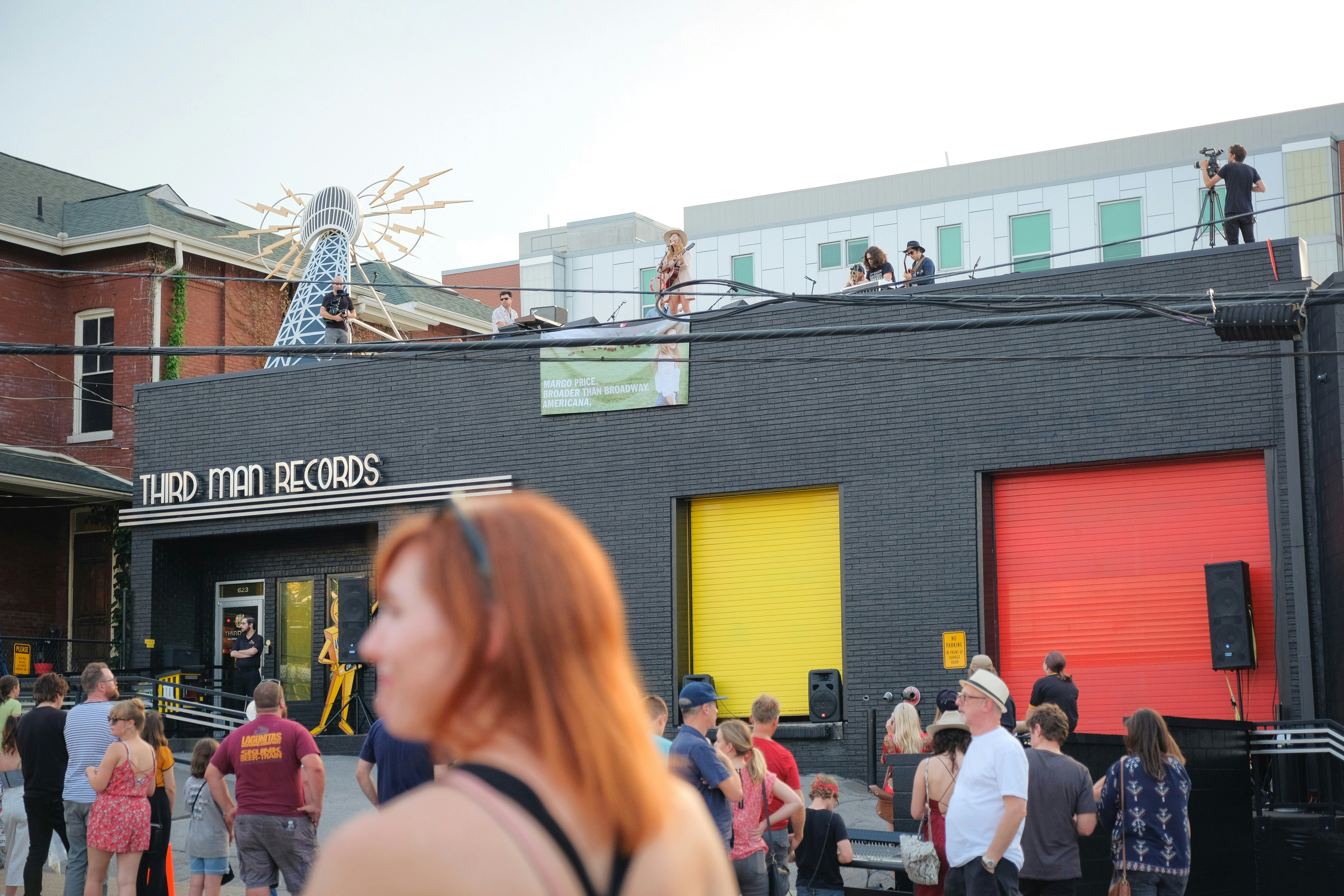 A red haired woman in a black tank top with black sunglasses stands in profile in front of a small crowd watching a rooftop concert on the black brick building of Third Man Records, with yellow and red brick insets in the facade. 