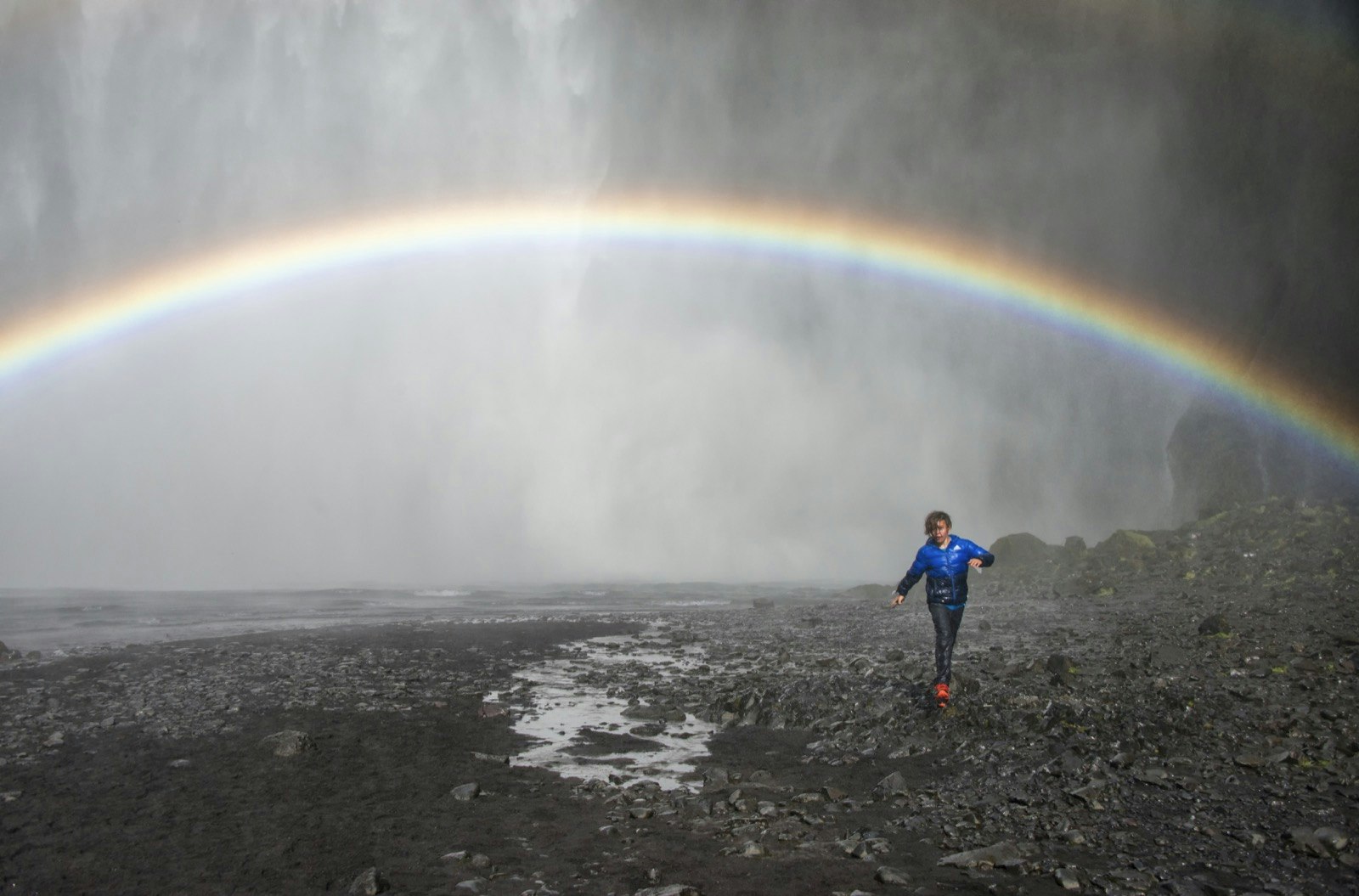 A young person walks under a rainbow created by the Skogafoss Waterfall