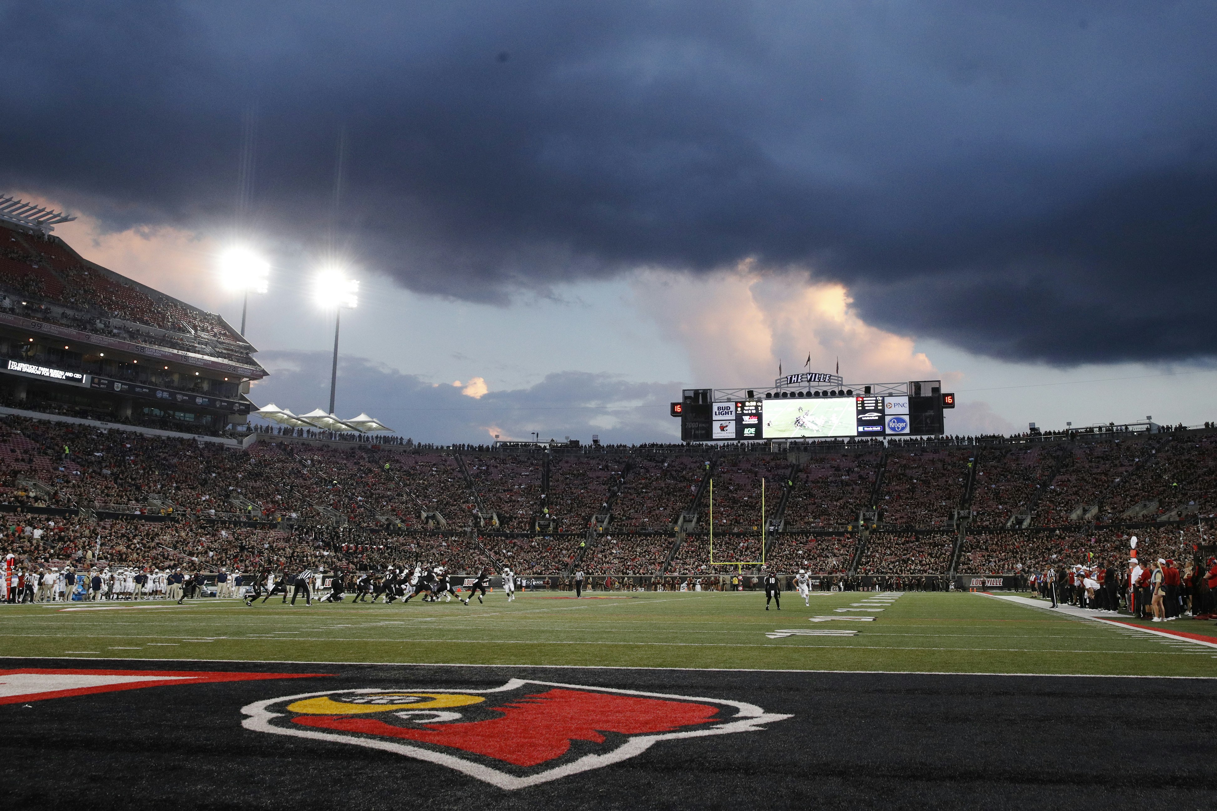 The foreground is taken up by the end zone of Cardinal Stadium, the turf painted black with a large red Cardinals logo appearing upside down tot the viewer. In the distance, players in black and white uniforms are in motion, and beyond them the stands full of fans. The sky over the stadium is blue with tall pink and grey cumulonimbus clouds overtaken by a huge black storm cloud hovering right over the stadium.