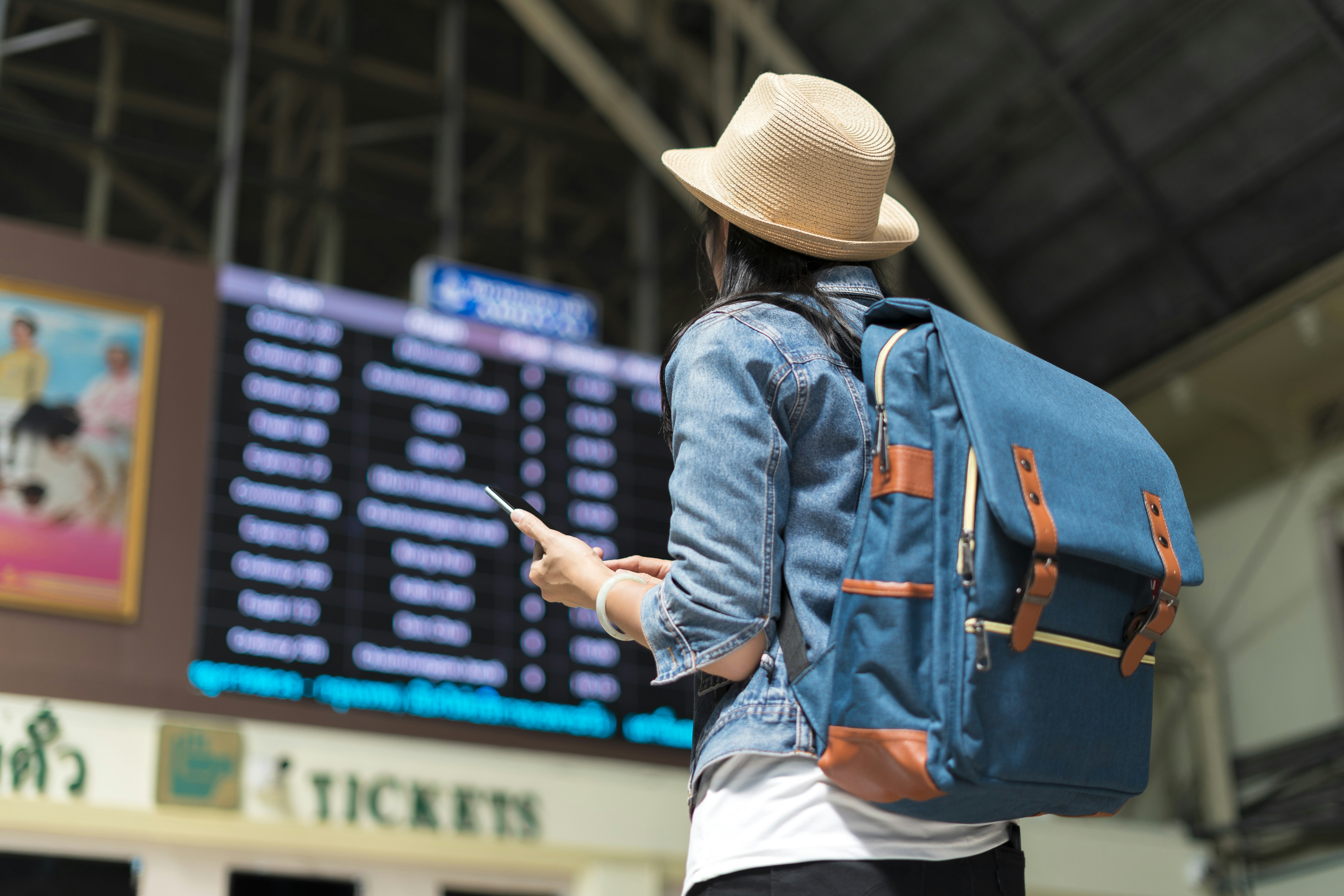 A young woman in a denim jacket and straw Panama hat gazes up at a departures and arrivals marquee in an airport