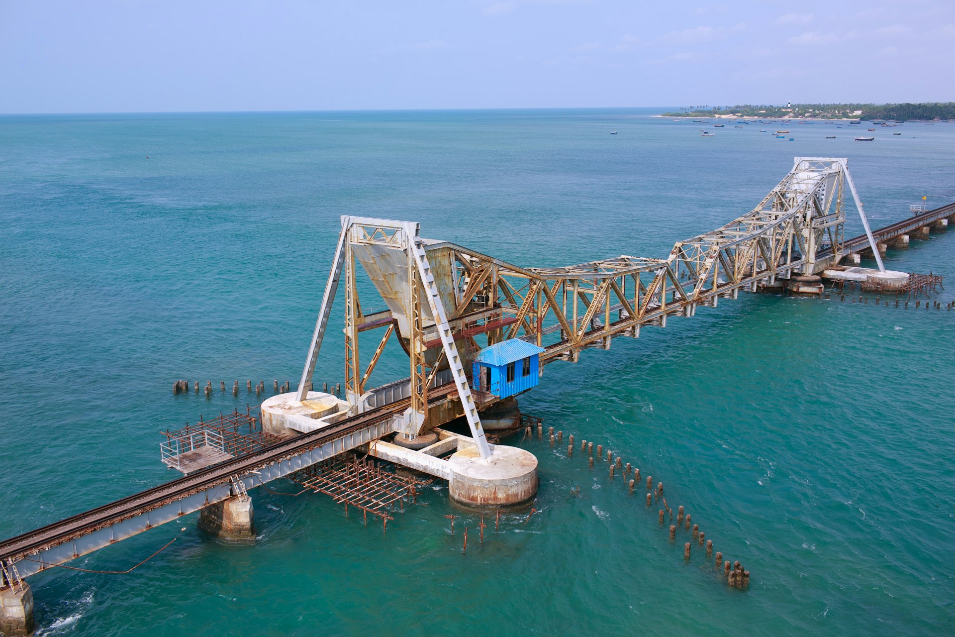 A white steel bridge tinged with rust diagonally spans this shot of the ocean between Pamban Island and mainland India. A bright blue control cabin sits on one side of the cantilevered Pamban Bridge, while an arc of wood buoys curves towards the viewer. In the background, small ships anchored offshore can be seen by the horizon 