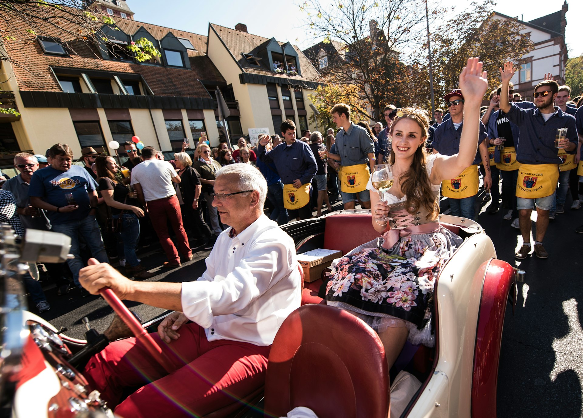 A young woman with long brown hair wearing a pink, blue, and light brown dirndl with a floral print on the skirt waves with her left hand as she holds a big stemmed glass of white wine in her right. She sits in a vintage white convertible with a red interior driven by a white-haired man in red pants a white silk shirt. Behind the Wine Princess are men in blue shirts with yellow aprons in formation as for a parade.