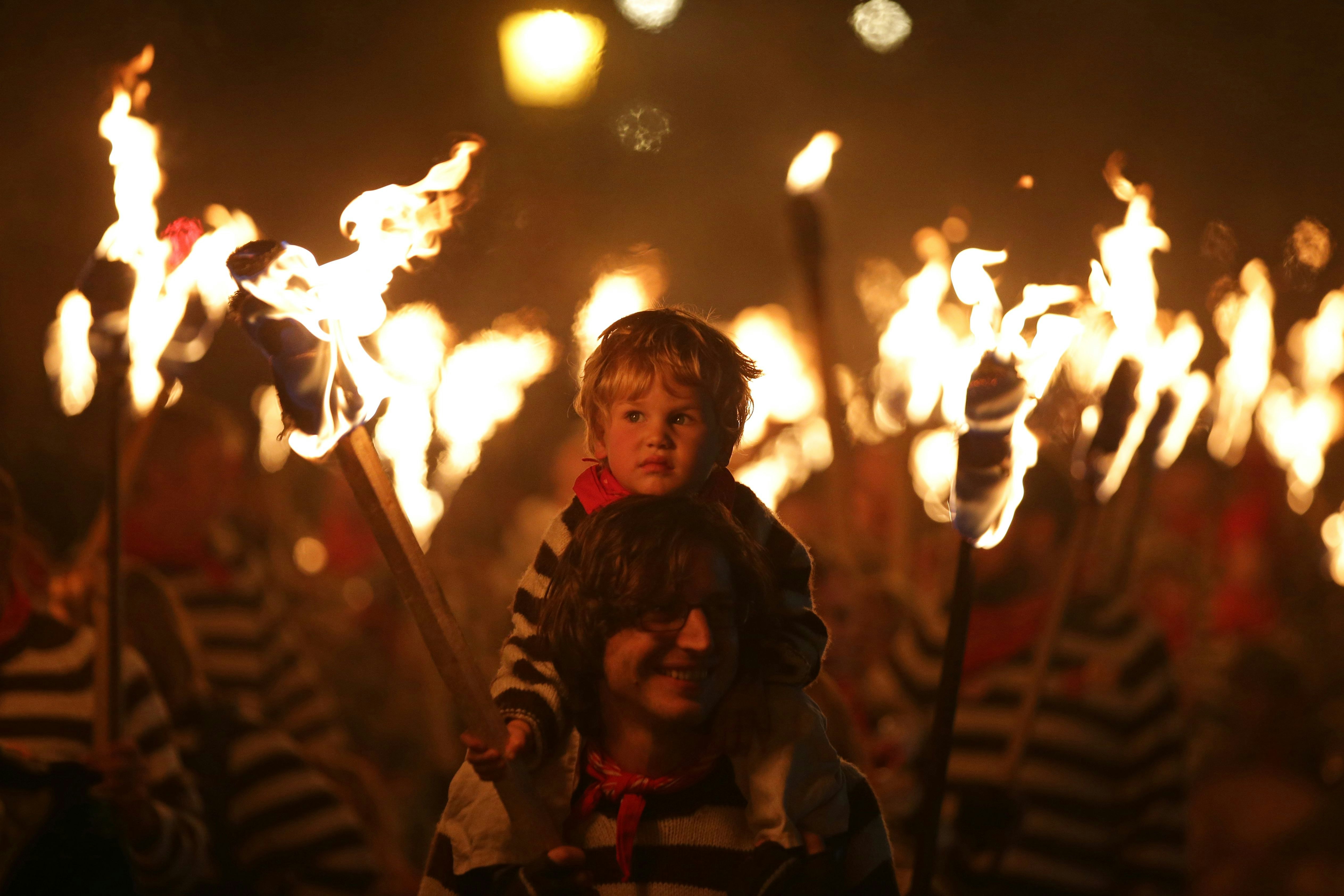 A child on an adult's shoulders holds a flaming torch. Everyone in the crowd is dressed in black and white stripes with a red neckerchief, and many are waving flaming torches