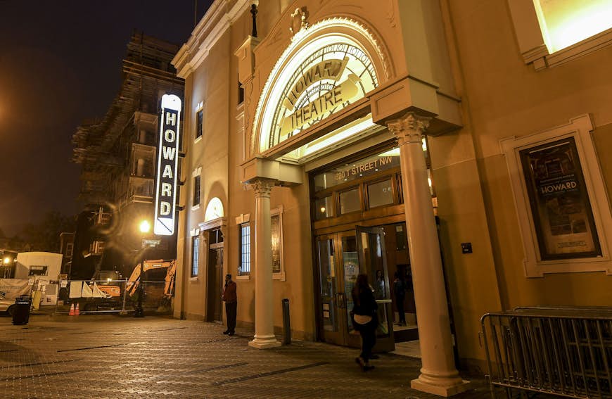   En nattlig bild av entrén till Howard Theatre.  En skylt är upplyst med namnet 'Howard' som löper vertikalt, och en kvinna går in i teatern mellan två kolumner i entrévägen.
