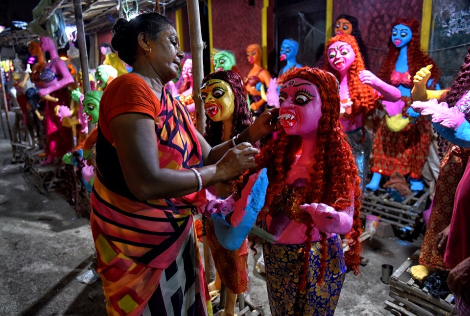A female artist in a colorful sari paints a bright pink clay statue of a female demon with long red curly hair and a blue and gold skirt. In the background are several other similar statues in a variety of bright primary colors.