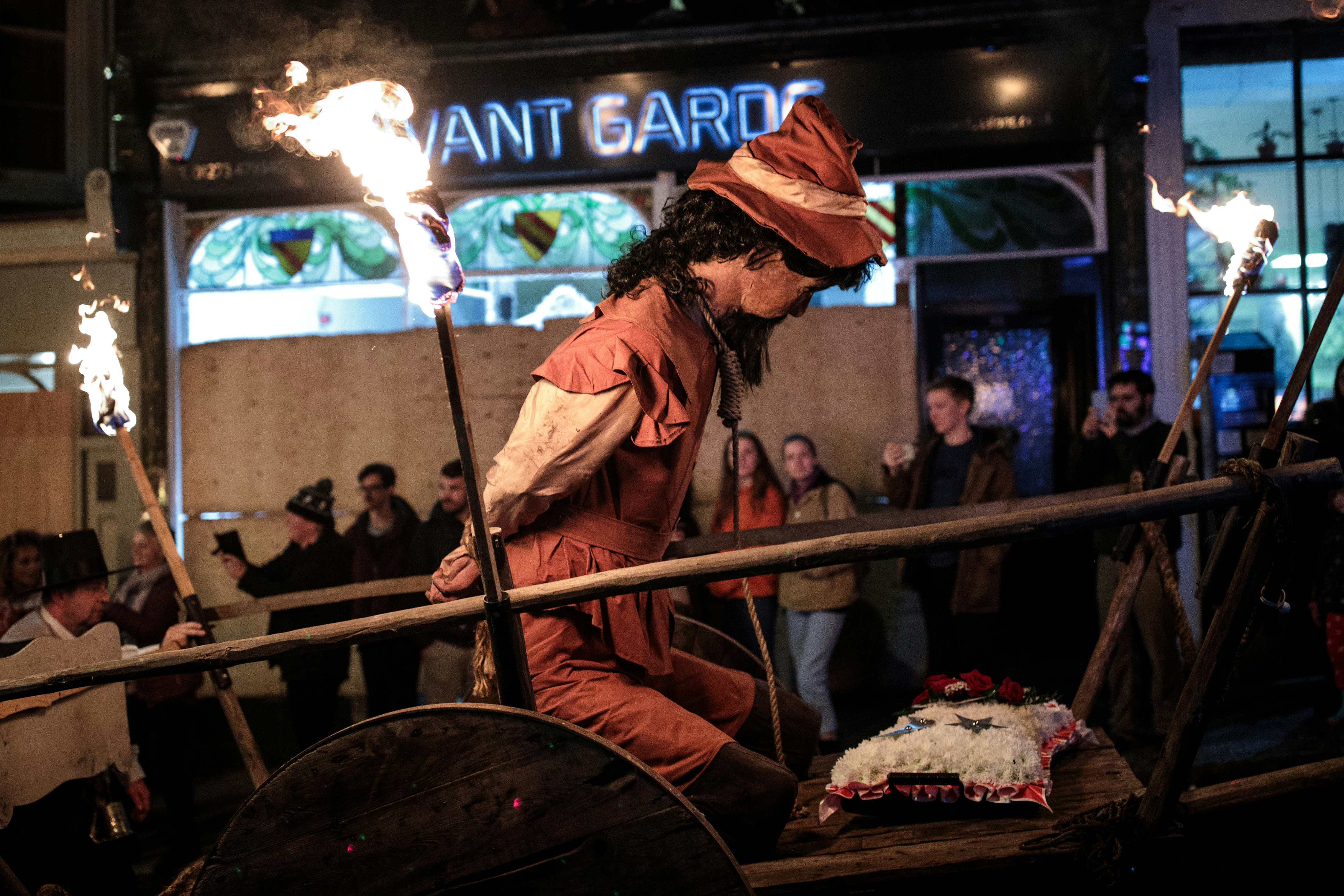 A kneeling effigy of Guy Fawkes sitting on a wooden cart is being pulled through the streets of Lewes. Fawkes is dressed in medieval garb and his hands are tied behind his back. Beyond the cart a shop with boarded-up windows is visible.