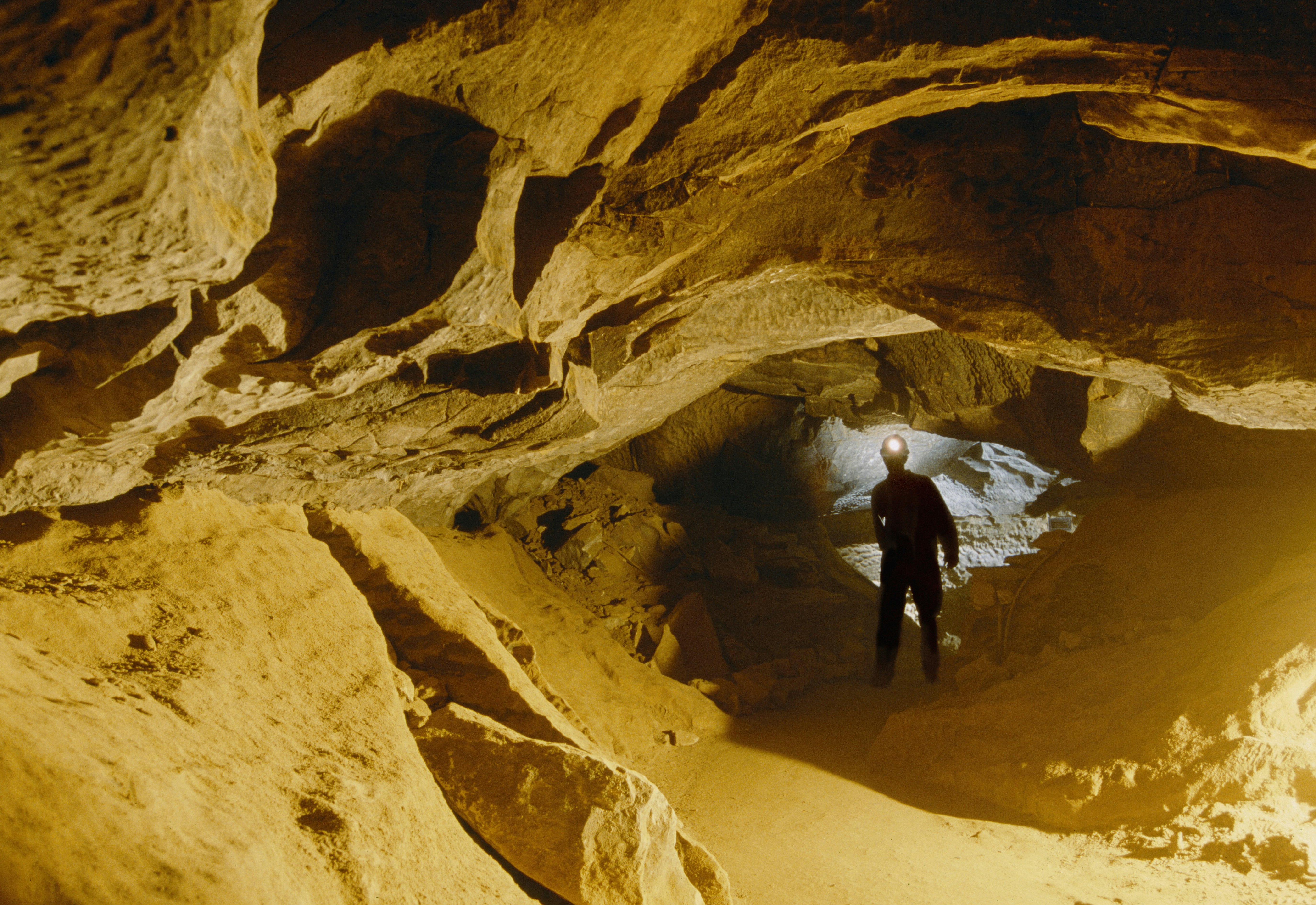 The silhouette of a man wearing a head torch can be seen walking through a narrow passageway in a cave system. The cave walls are a sandy-yellow color.