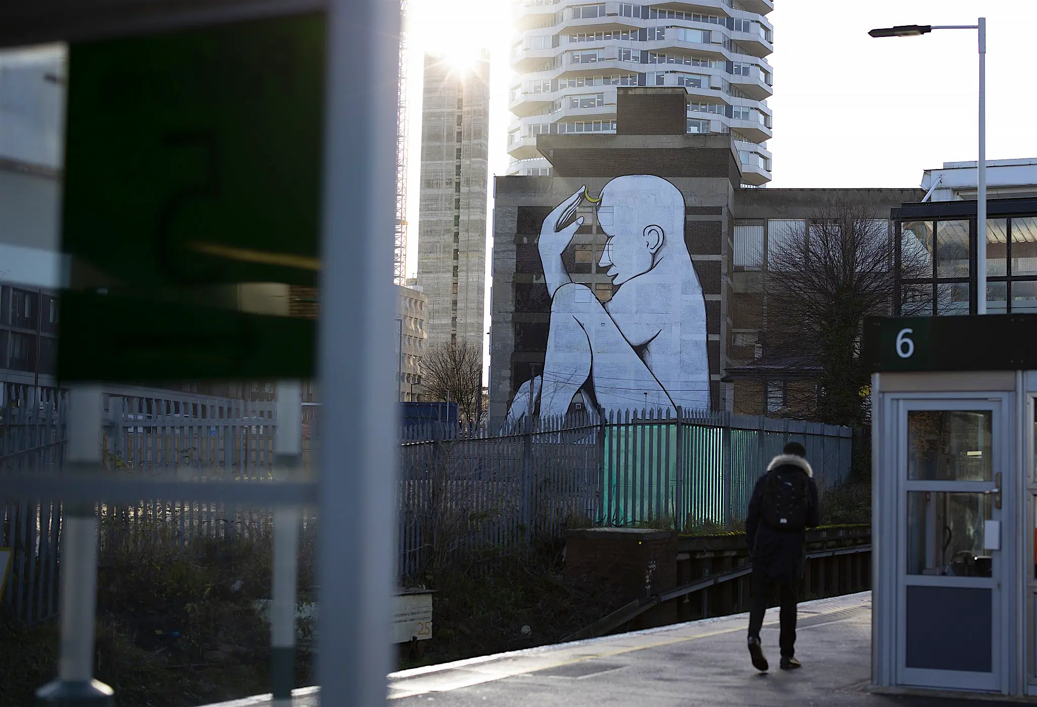 Croydon street art A teenager walking along a train station platform, with tower blocks visible beyond; on one of them is emblazoned a huge mural of a naked seated white figure.