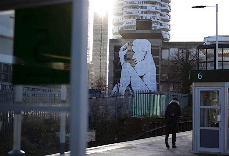 Croydon street art A teenager walking along a train station platform, with tower blocks visible beyond; on one of them is emblazoned a huge mural of a naked seated white figure.