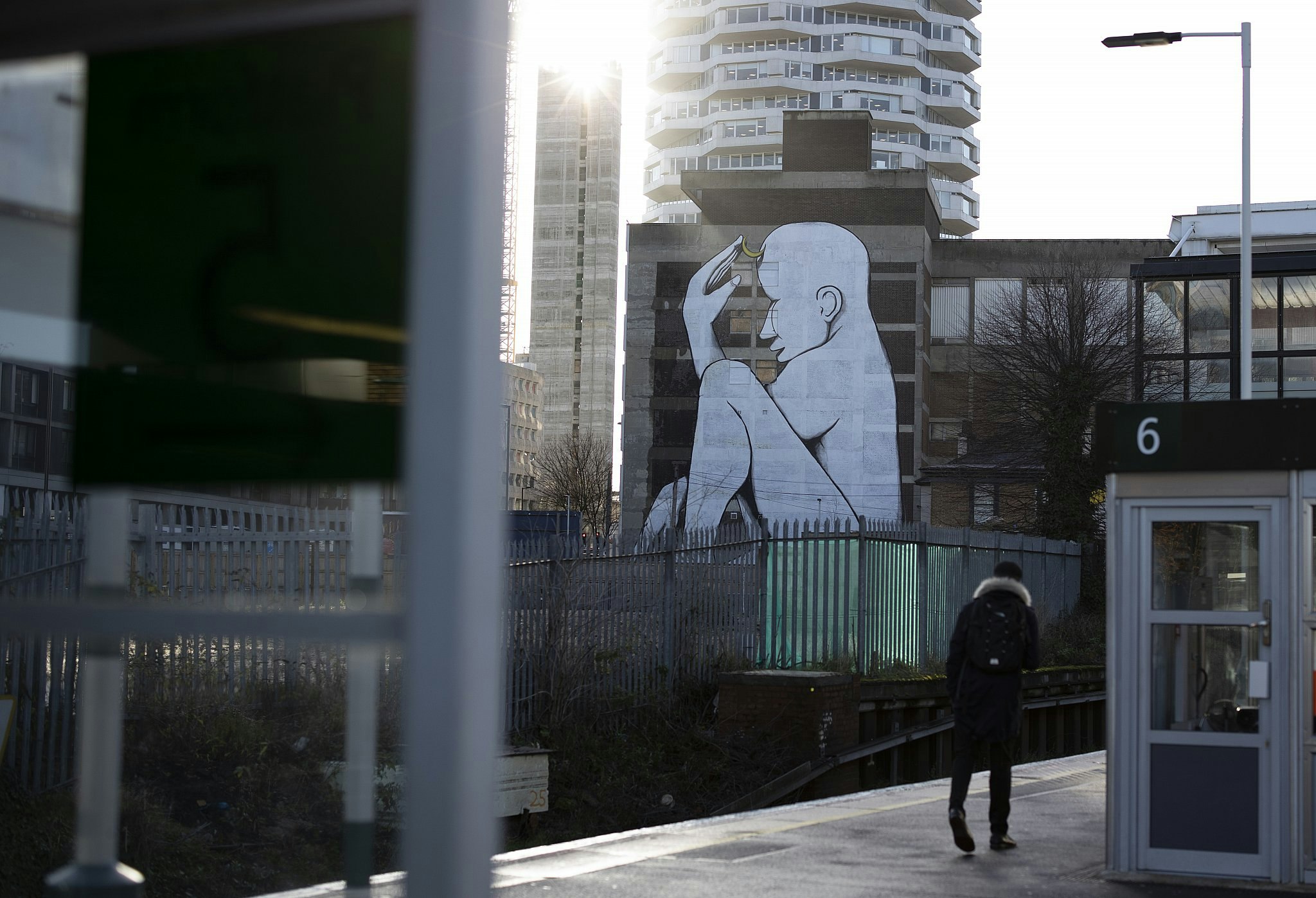 A teenager walking along a train station platform, with tower blocks visible beyond; on one of them is emblazoned a huge mural of a naked seated white figure. 