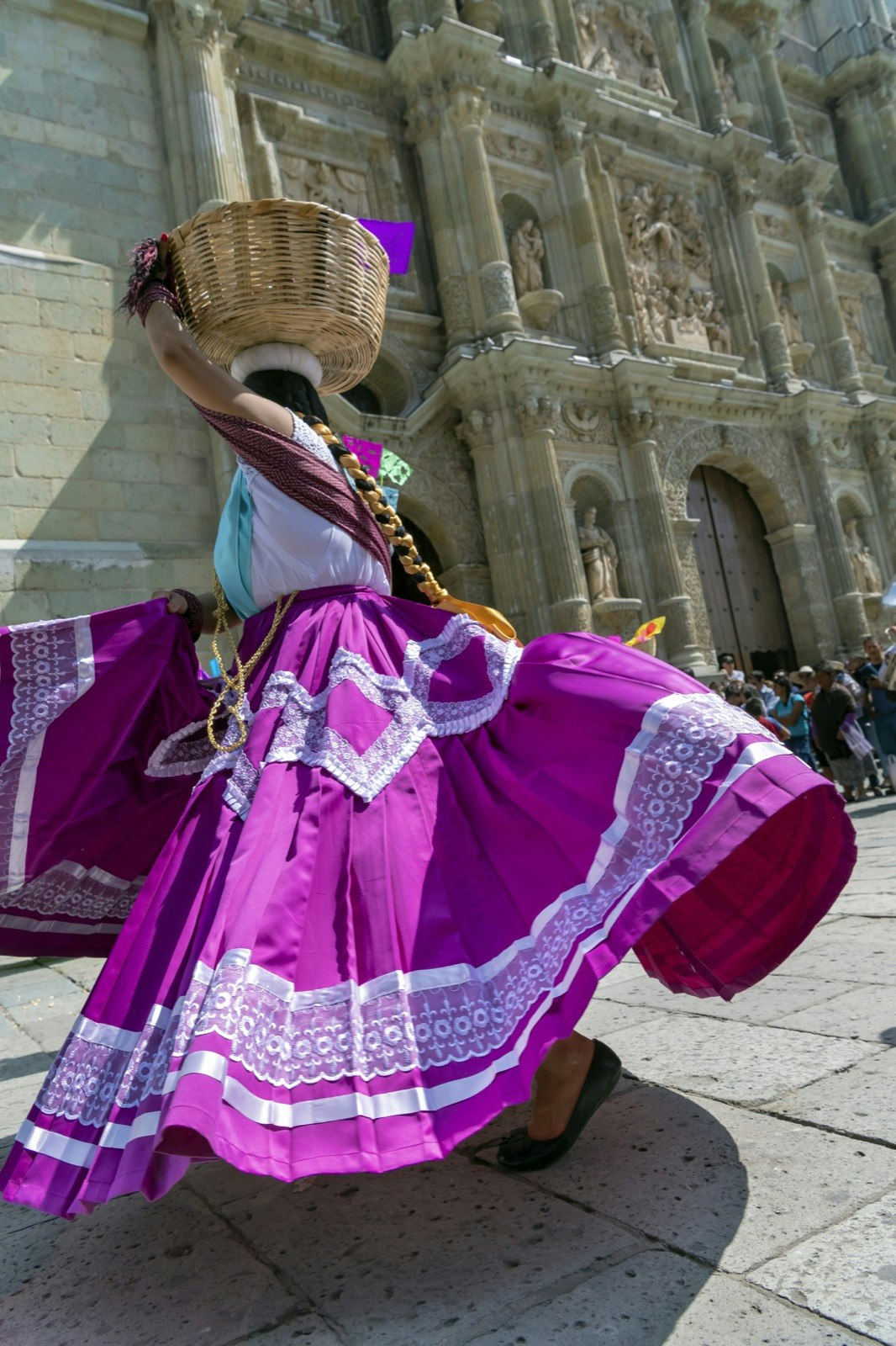 A woman holds a woven basket on her head as she spins in her vibrant purple and white traditional flared skirt in the middle of a town square; Dia de Muertos