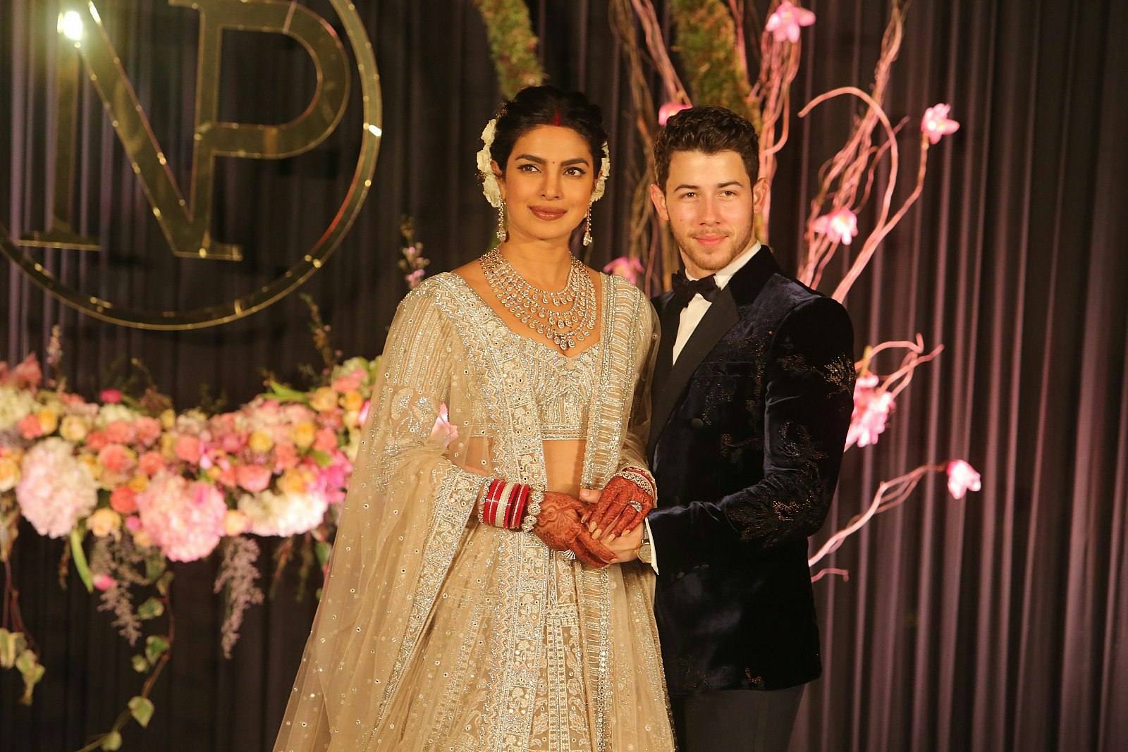 Priyanka Chopra and Nick Jonas at their wedding reception; she is wearing an elaborately beaded sari and necklace and he is wearing a dark velvet tuxedo. 