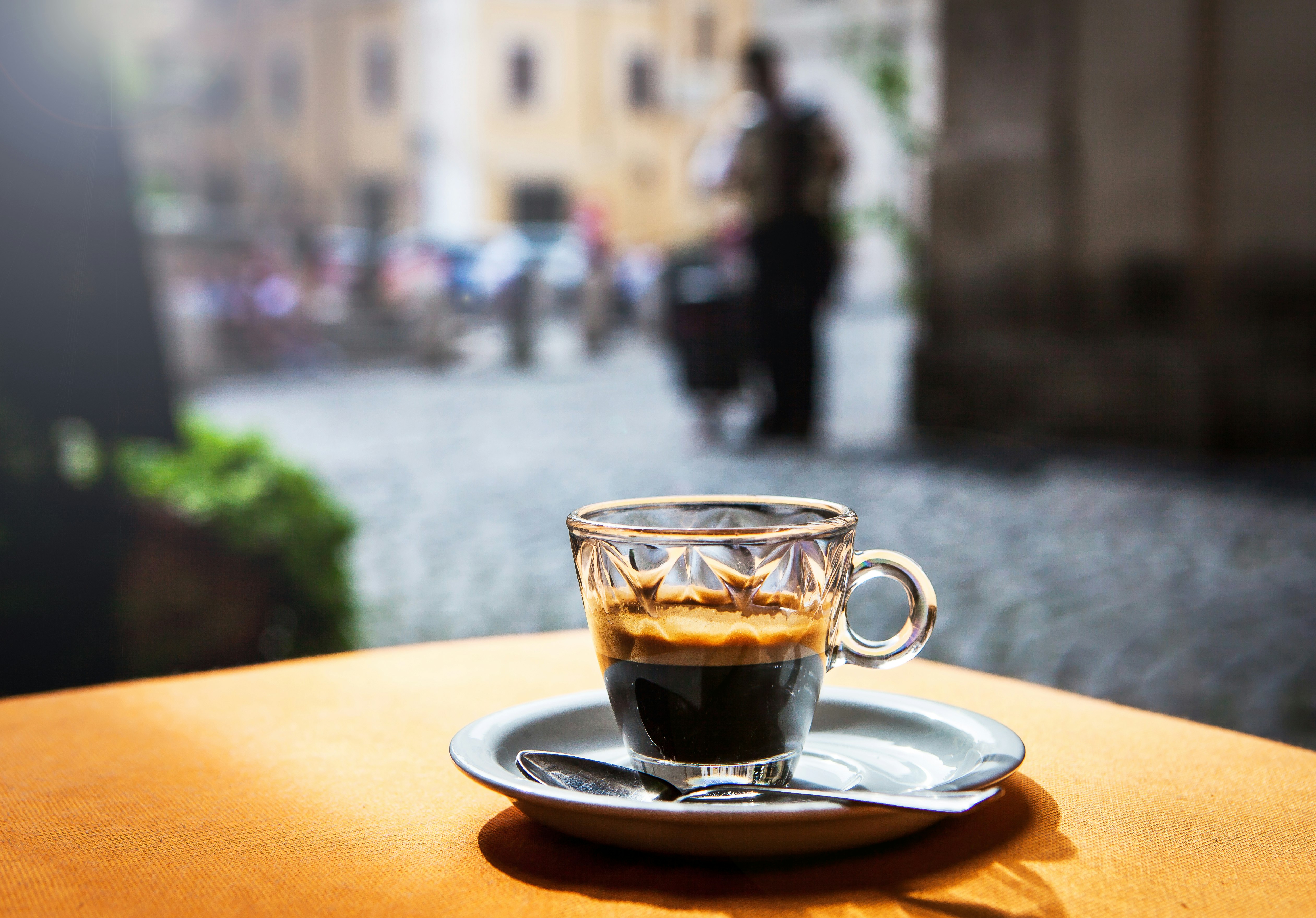 An espresso in a decorative glass cup sitting on a saucer on a table outside. The street scene in the background is in soft focus.