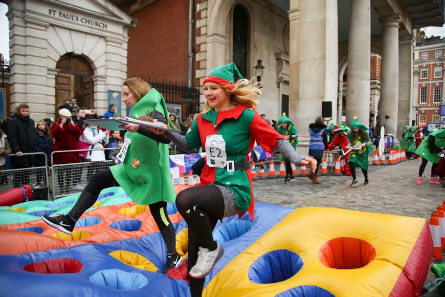 Two women dressed in costume - one as an elf, one as a Christmas tree - run over an inflatable with holes while carrying plates with Christmas pudding