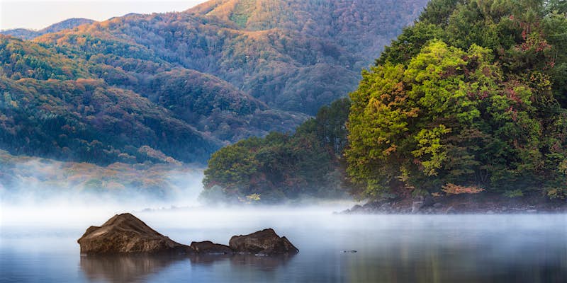 A foggy morning at Hibara lake in Fukushima. The lake's still waters are shrouded in fog, from which three small boulders emerge. Around the lake are hills covered in dense forest.