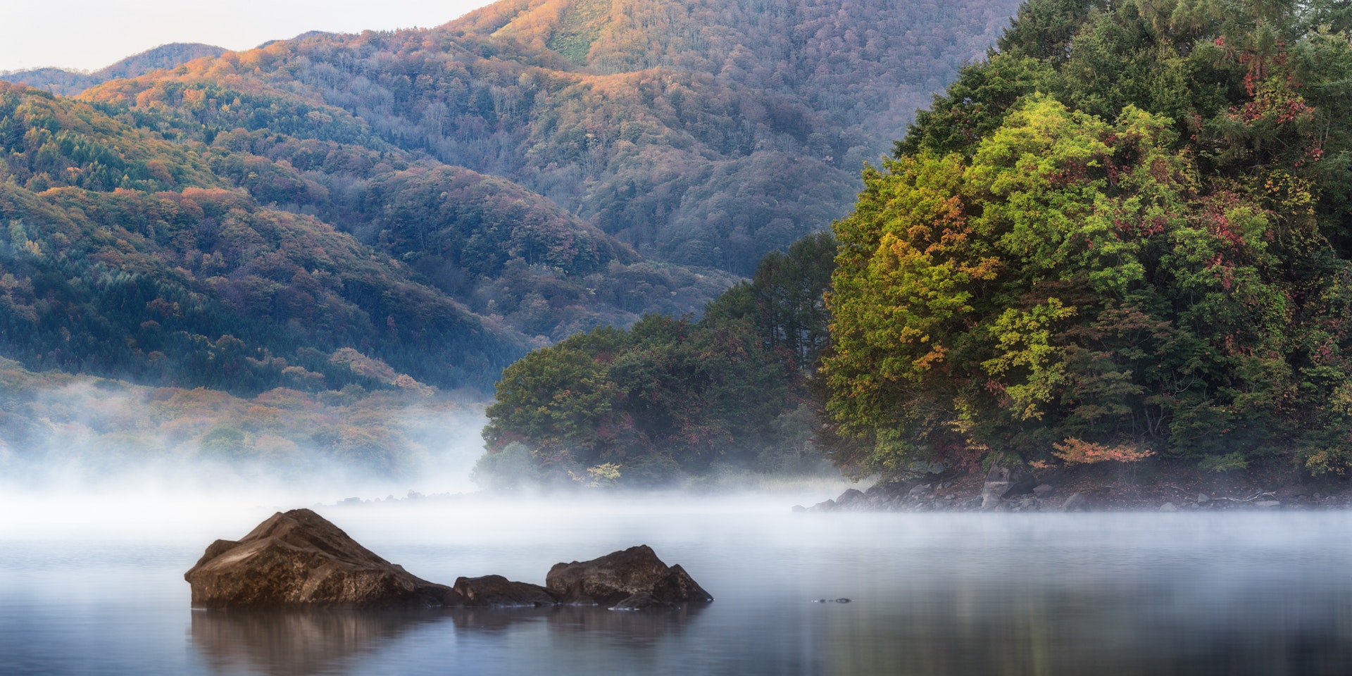 A foggy morning at Hibara lake in Fukushima. The lake's still waters are shrouded in fog, from which three small boulders emerge. Around the lake are hills covered in dense forest.