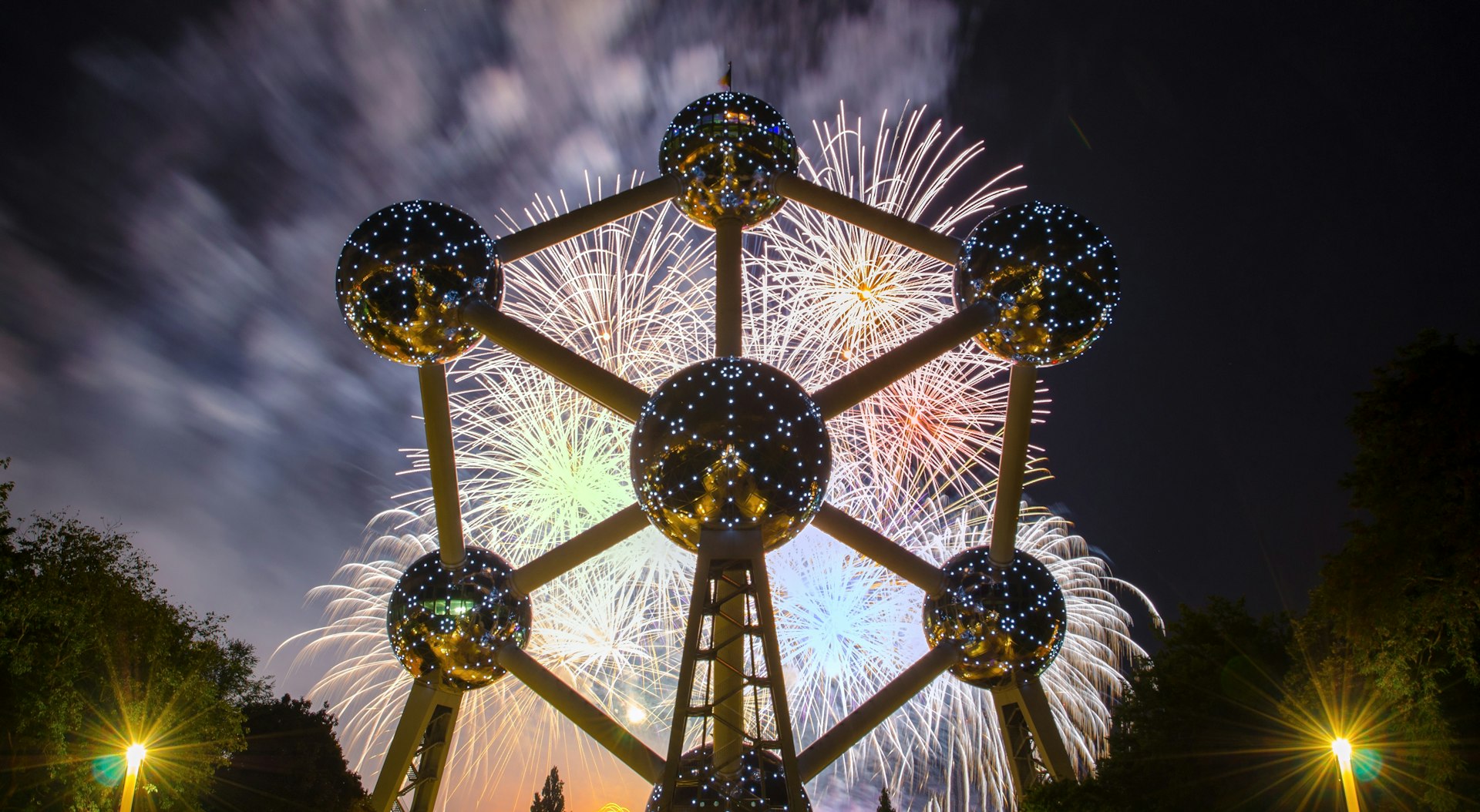 A large metal sculpture in the shape of an atom with six big silver balls connected to a larger, central sphere by metal rods is lit by both a series of lights arranged in similar atomic patterns on the surface of each sphere and by a bright fireworks display behind the Atomium against a deep cobalt sky