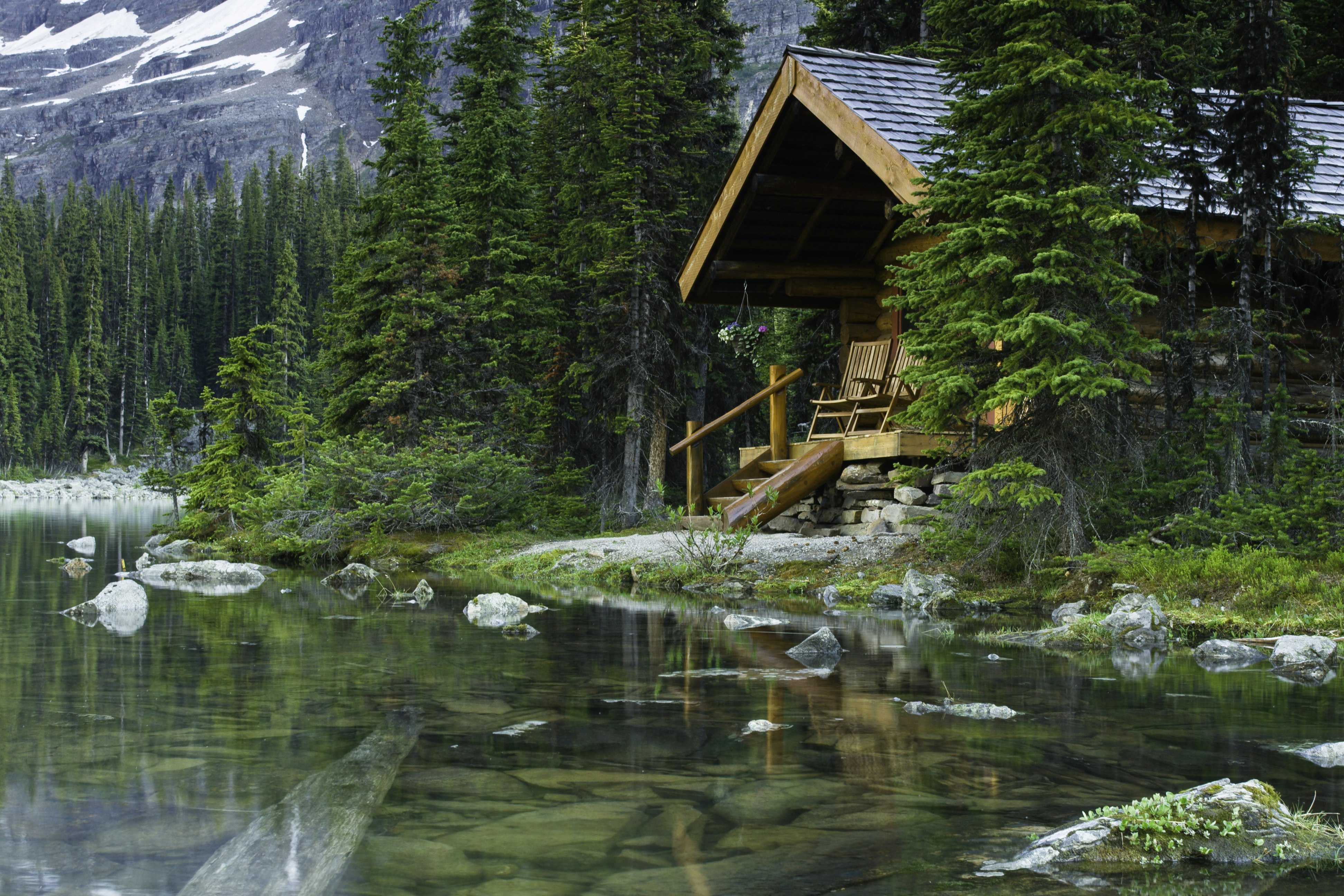 A log cabin set within evergreen trees on the rocky shore of a peaceful lake.