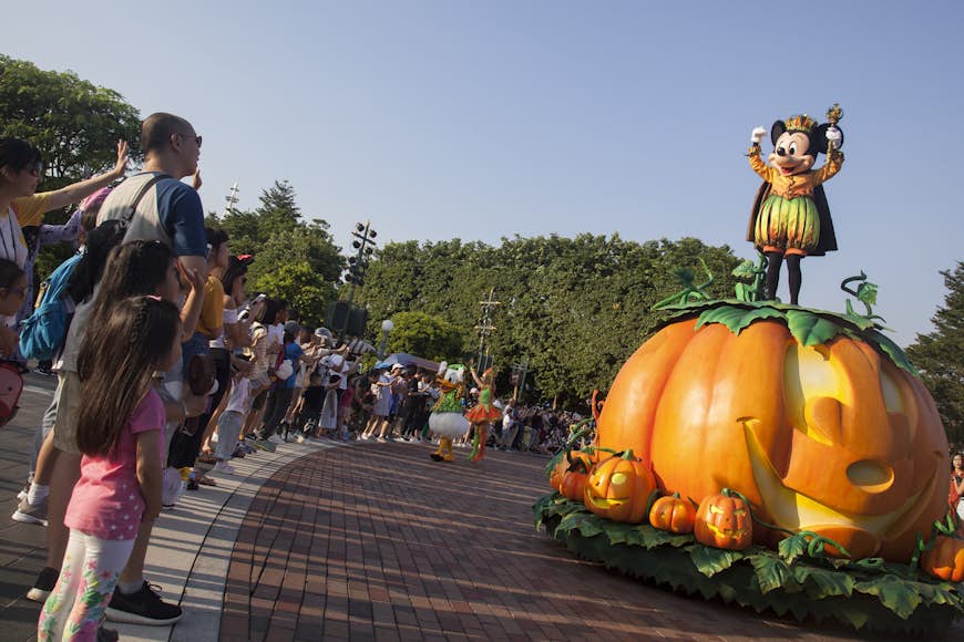 Tourists wave to Mickey Mouse, who stands proudly atop a giant pumpkin as part of the Hong Kong Disneyland Halloween Parade.