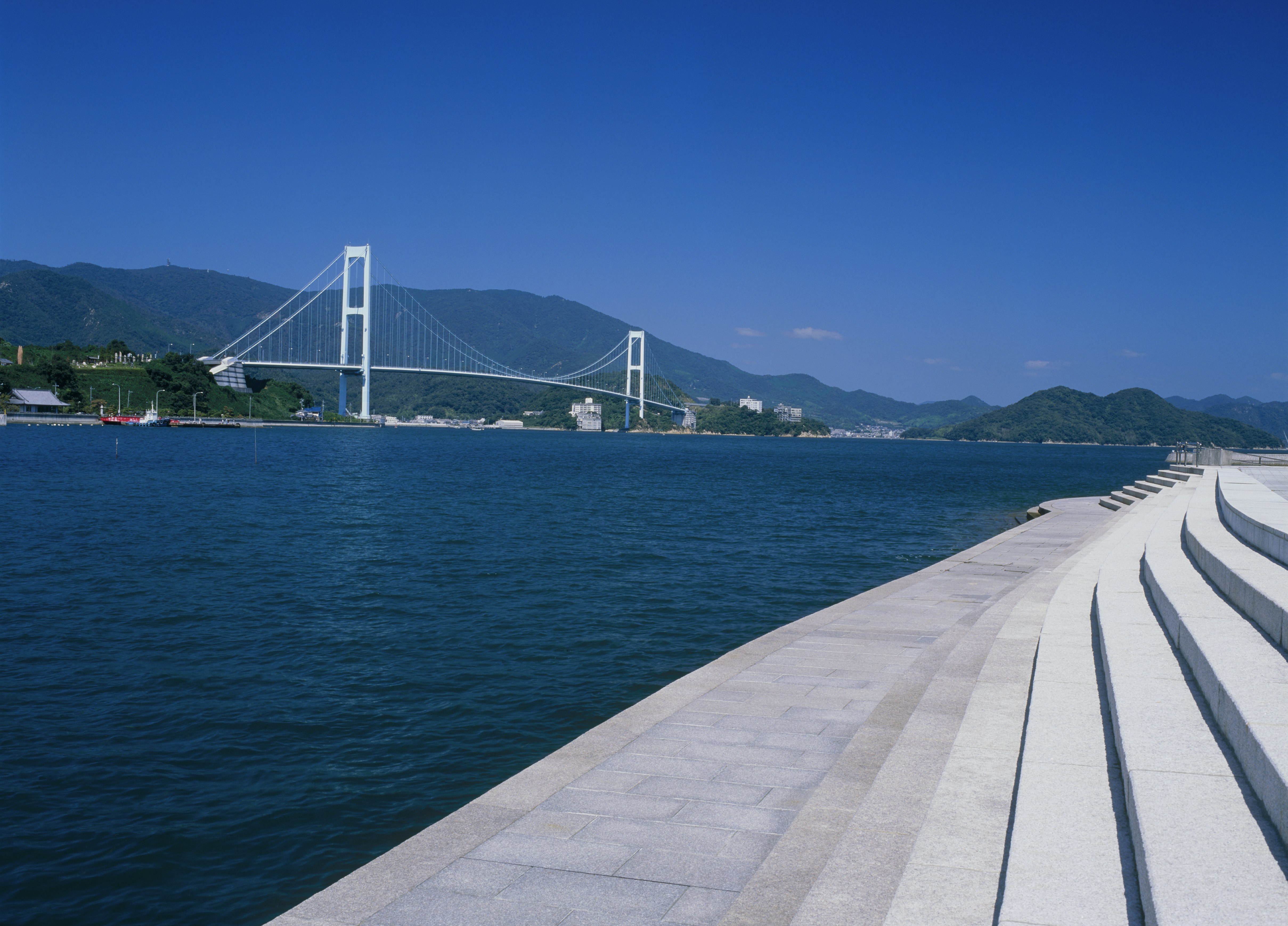 The Akinaada Bridge is a white suspension bridge with simple, modernist forms. It's shot from a series of concrete steps in Kure, Hiroshima that go right down to the deep, denim-blue water. In the distance at the foot of the bridge is a red ship, thin white streetlights, and some small grey buildings. On either end and behind it are tall green and blue mountains. The sky is a bright, deeply saturated blue.