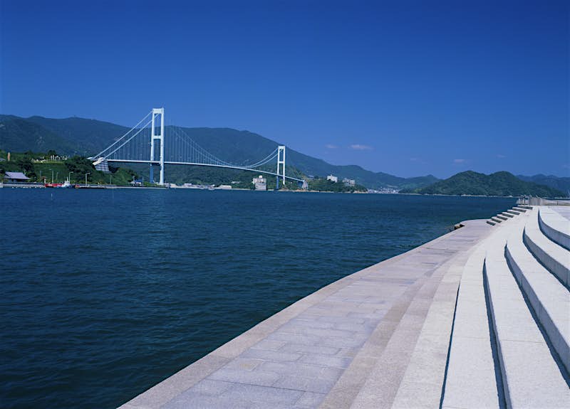 Akinaada-bridge.jpg The Akinaada Bridge is a white suspension bridge with simple, modernist forms. It's shot from a series of concrete steps in Kure, Hiroshima that go right down to the deep, denim-blue water. In the distance at the foot of the bridge is a red ship, thin white streetlights, and some small grey buildings. On either end and behind it are tall green and blue mountains. The sky is a bright, deeply saturated blue.