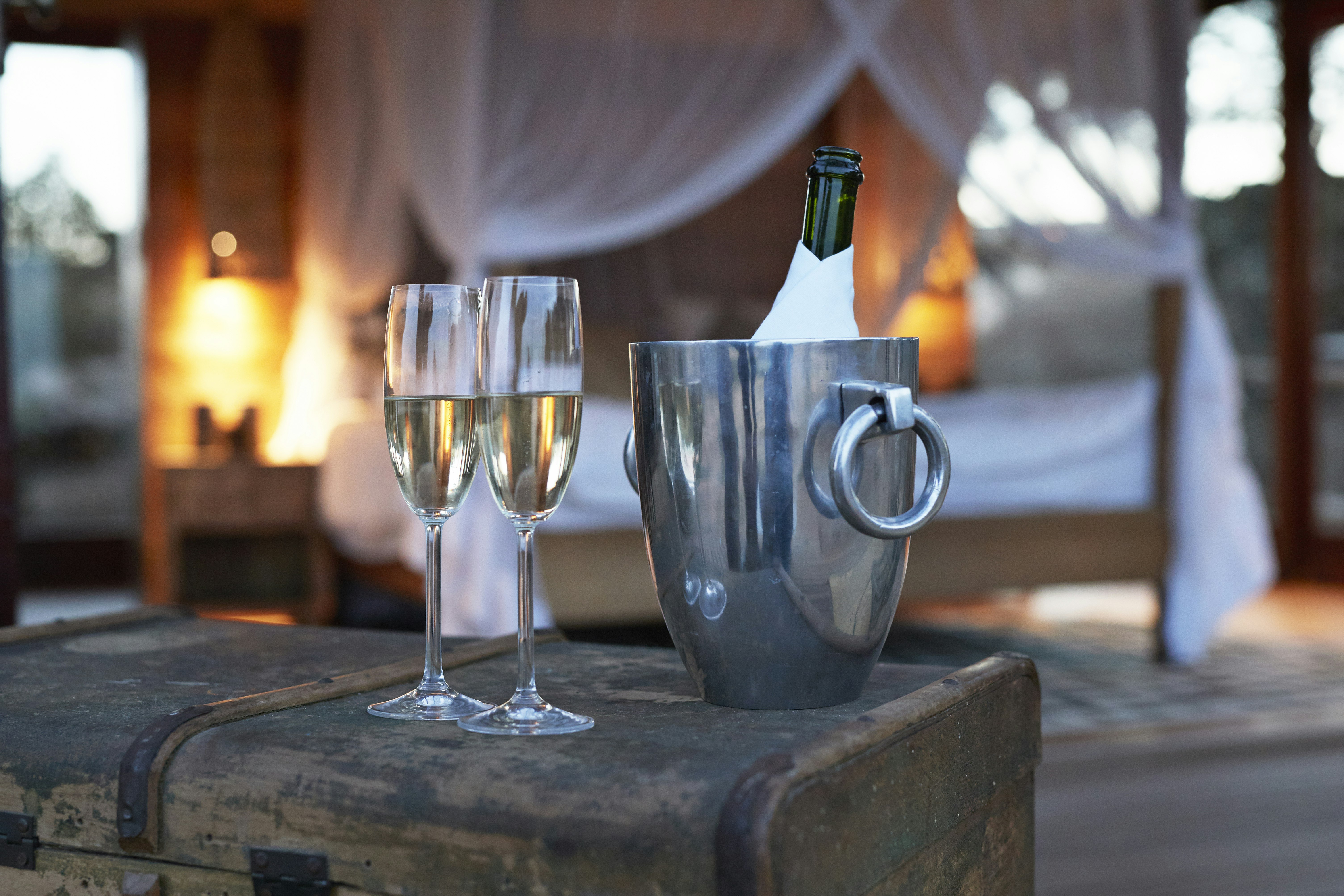 A bottle of champagne in a silver ice container sits next to two glasses of bubbly; in the background is a roaring fire.