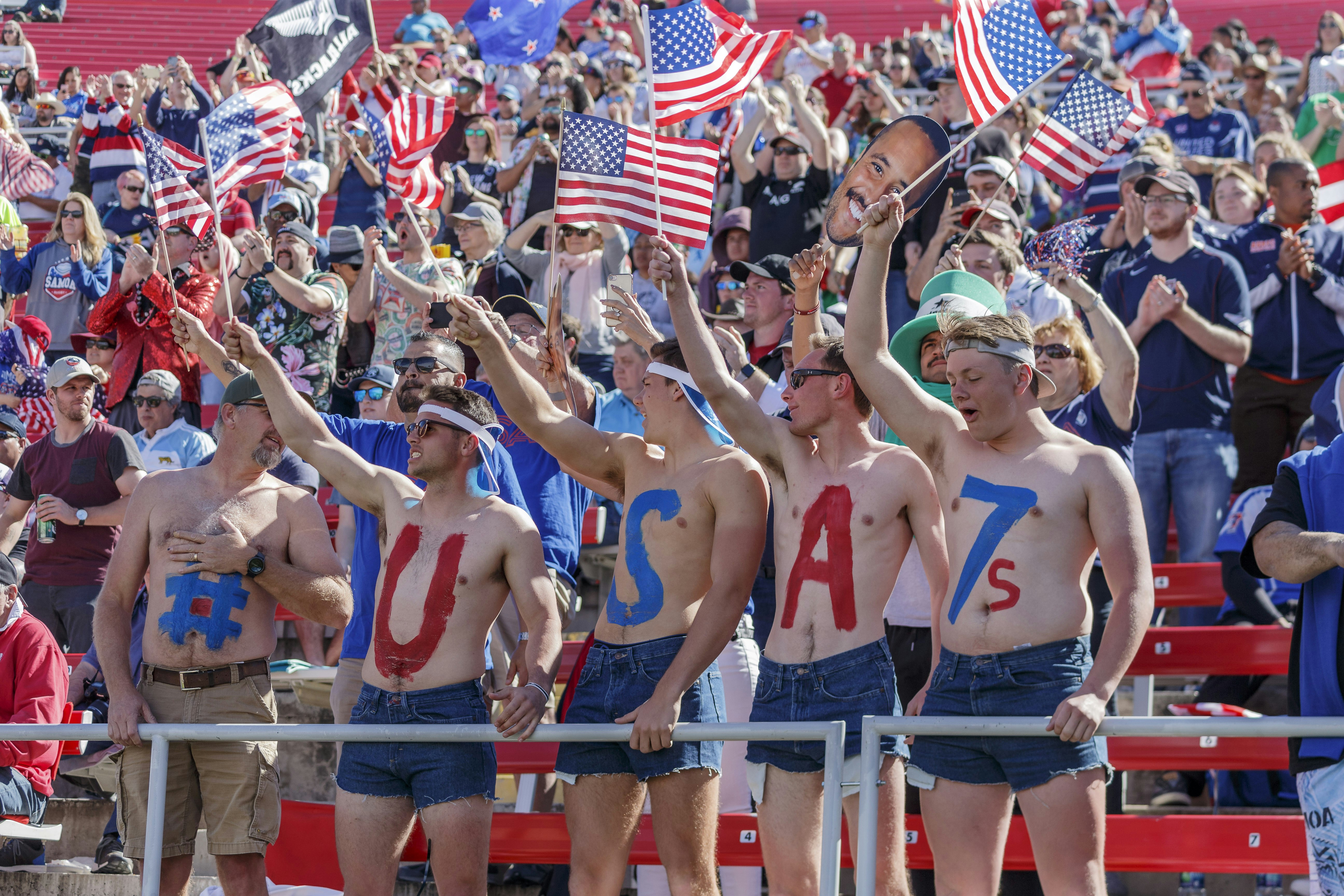 Five USA fans stand topless in the stands at the USA Rugby Sevens tournament in Las Vegas. Each of the five has a letter or symbol on their chest, which spells out: '#USA7s'. Behind them other fans clap and wave flags.