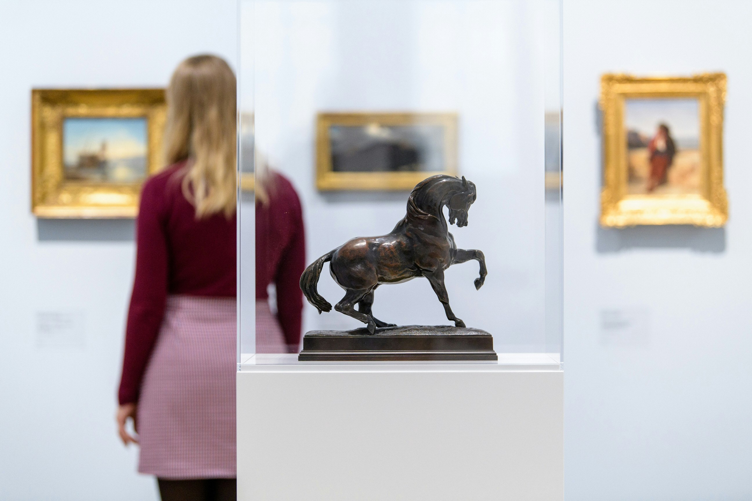 Inside a gallery, a woman is looking at one of three paintings in gold frames, hanging on a white wall. She is in soft focus, while a miniature bronze sculpture of a horse inside a glass case is in the foreground.