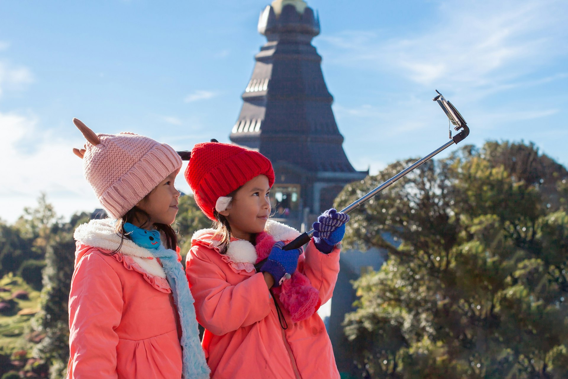 Two young children, dressed in coats and furry hats, take a selfie together in Chiang Mai, with a temple stupa visible in the background.
