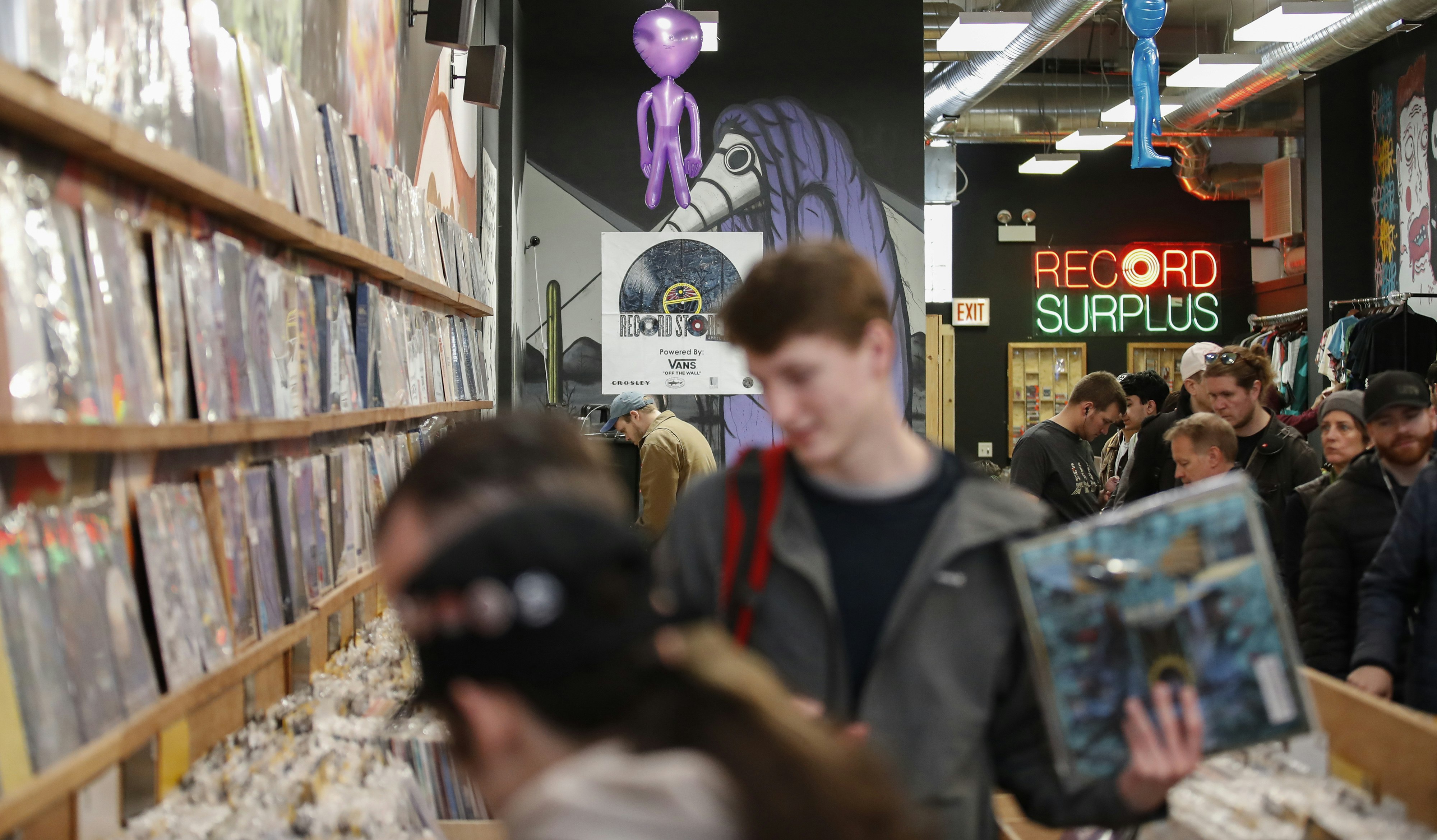 Shoppers sort through bins of albums at Shuga Records. In the center of the image is a young man with red hair in a grey hoodie with a red backpack holding a blue album. In focus behind him is a purple mural, a purple inflatable alien toy, and a neon sign in red and green reading "Record Surplus" where the O in Record looks like an album 