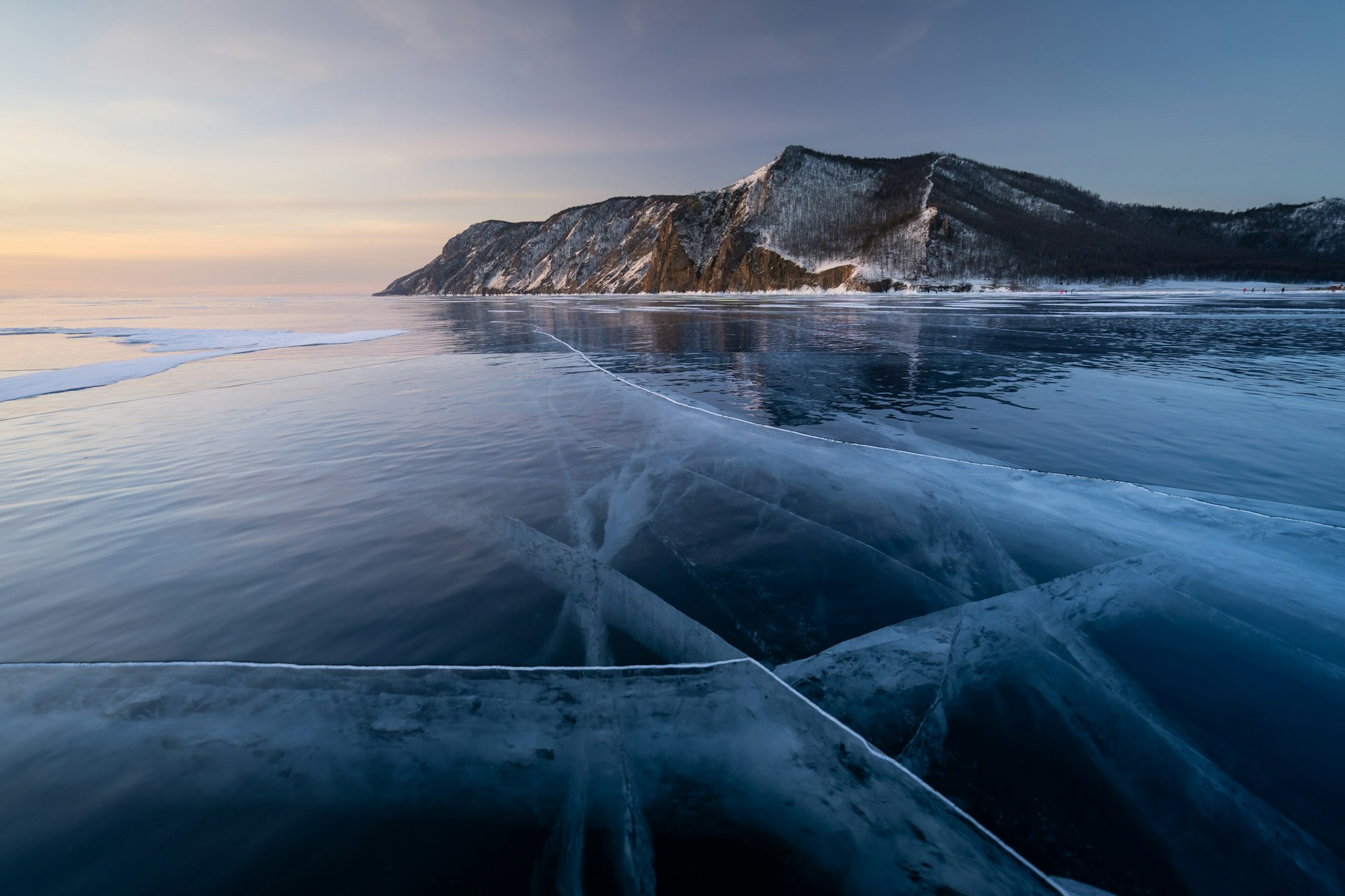 The sun rises over a steep rocky cliff standing up from the shoreline of the partially frozen Lake Baikal.