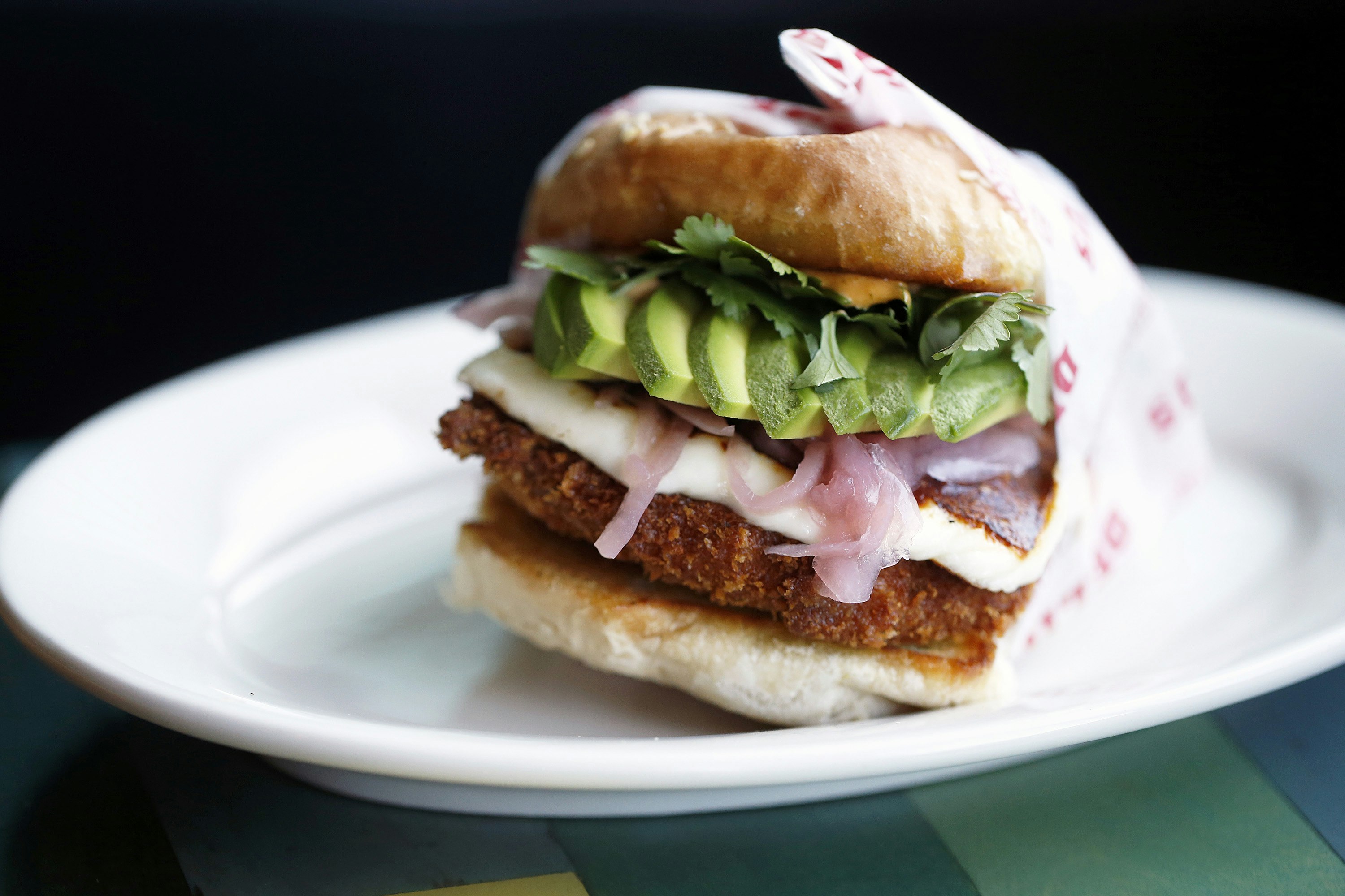 A cemita sandwich sits on a white plate. The bun is filled with breaded, deep-fried meat, onions, lettuce, cheese and avocado.