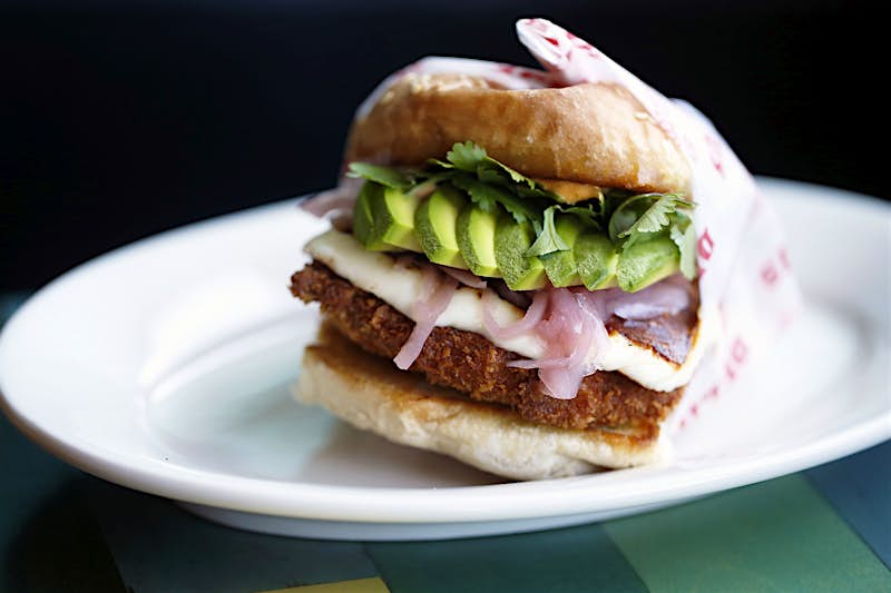 A cemita sandwich sit on a white plate. The bun is filled with breaded, deep-fried meat, onions, lettuce, cheese and avocado.