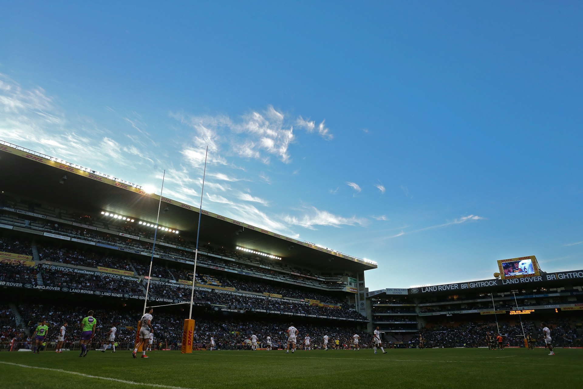 A low-angled view of the Newlands Stadium rugby pitch. A team is out on the field, waiting for the opposition to kick a penalty, while in the background a four-tiered stand is full of supporters.