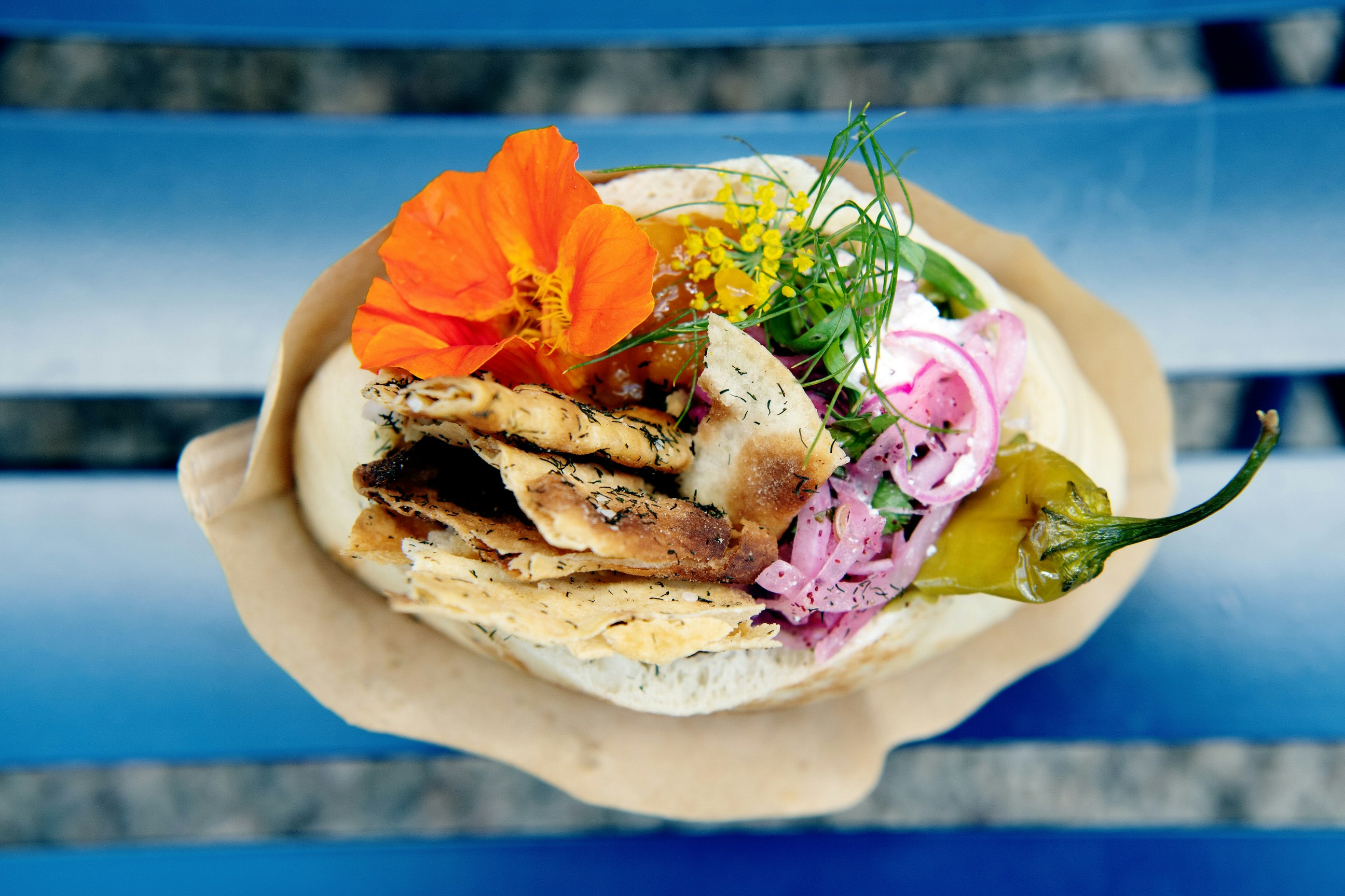 A close-up shot of a filled pita bread. Some of the  ingredients, such as red onion and a jalapeno can be seen, and it's decorated with an orange flower.