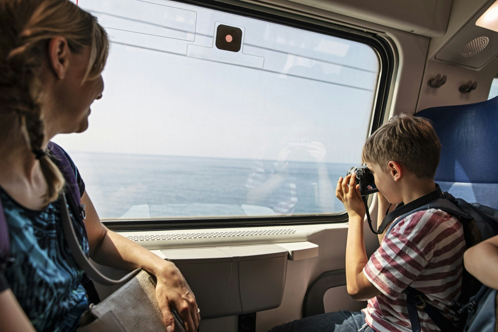 a mother and her son ride a train, the boy takes a photo out of the window while the mother smiles, they are in Italy