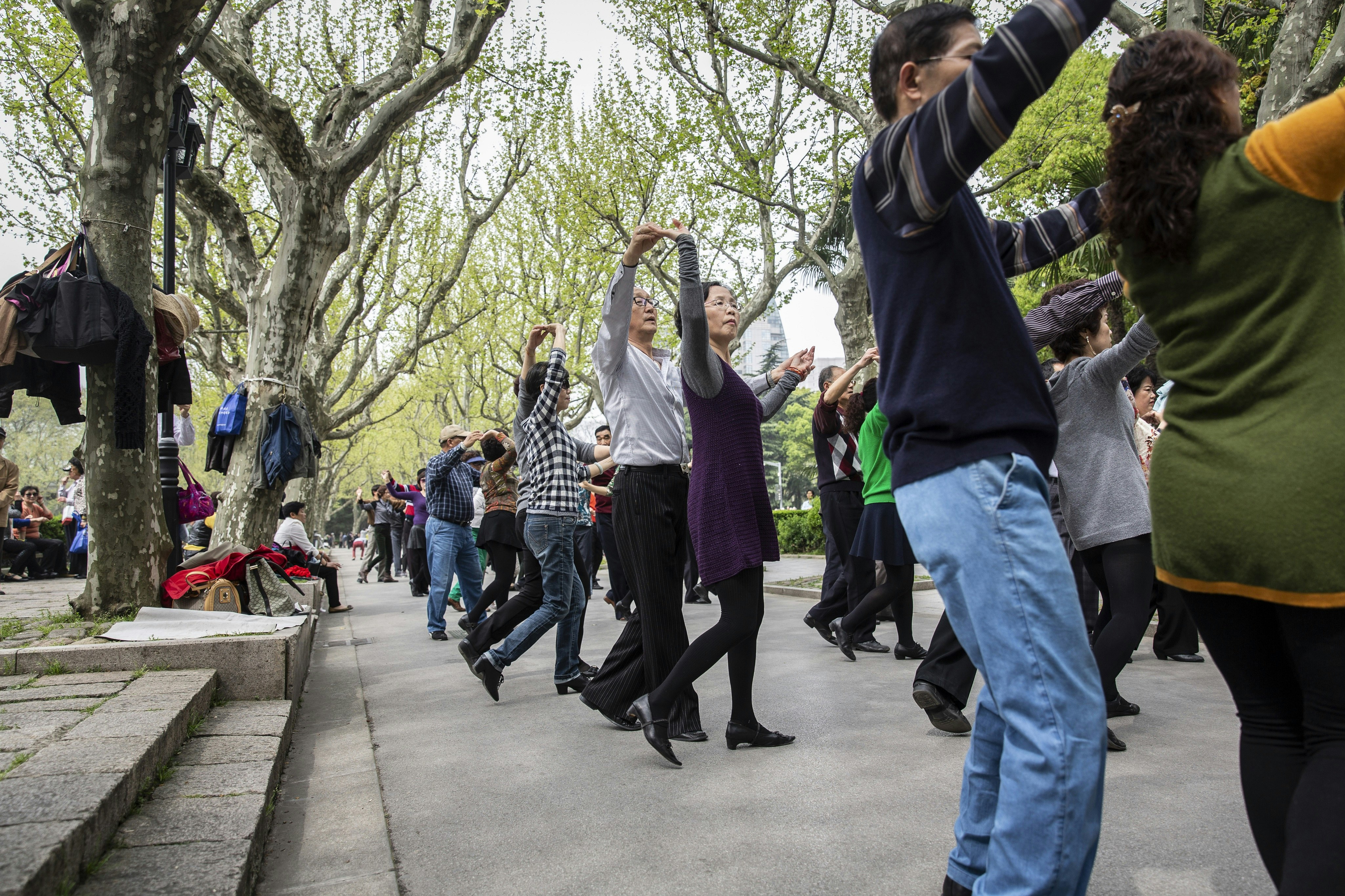 Par dansare i en lång rad spankulerar sina grejer på en betongdel av Fuxing Park.  Stigen är omgiven av träd och andra människor sitter på trappan för att titta på.