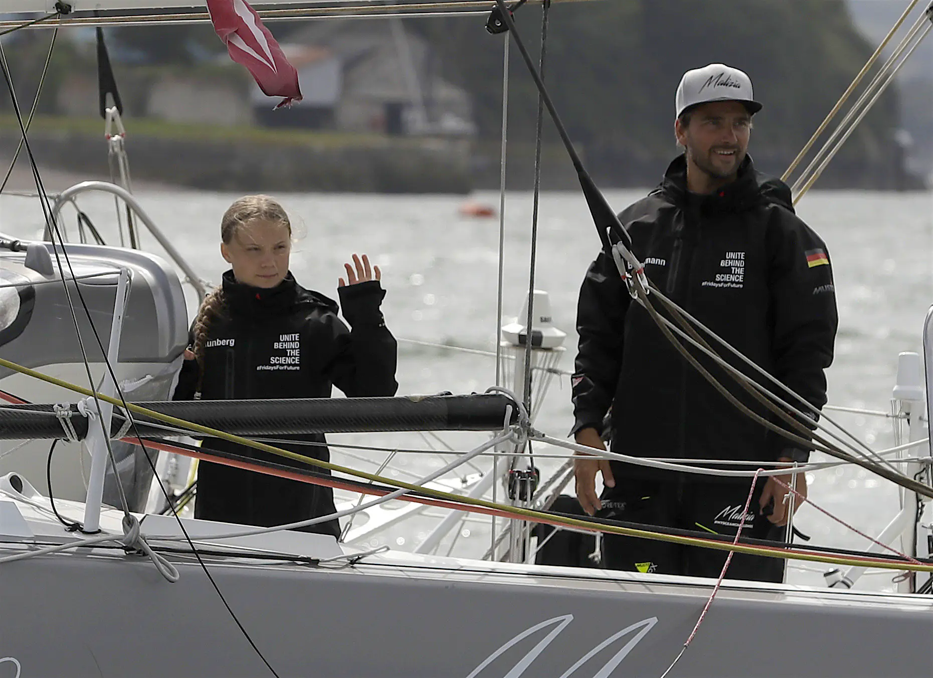 greta-thunberg-climate-change-activist-carbon-offset.jpg Climate activist Greta Thunberg stands on a solar powered sailboat ahead of her voyage to the 2019 United Nations Climate Action Summit. She wears a black windbreaker with the words United Behind the Science printed in white. She stands next to a man wearing a similar jacket, with the German flag on the left sleeve and a white ball cap on his head.