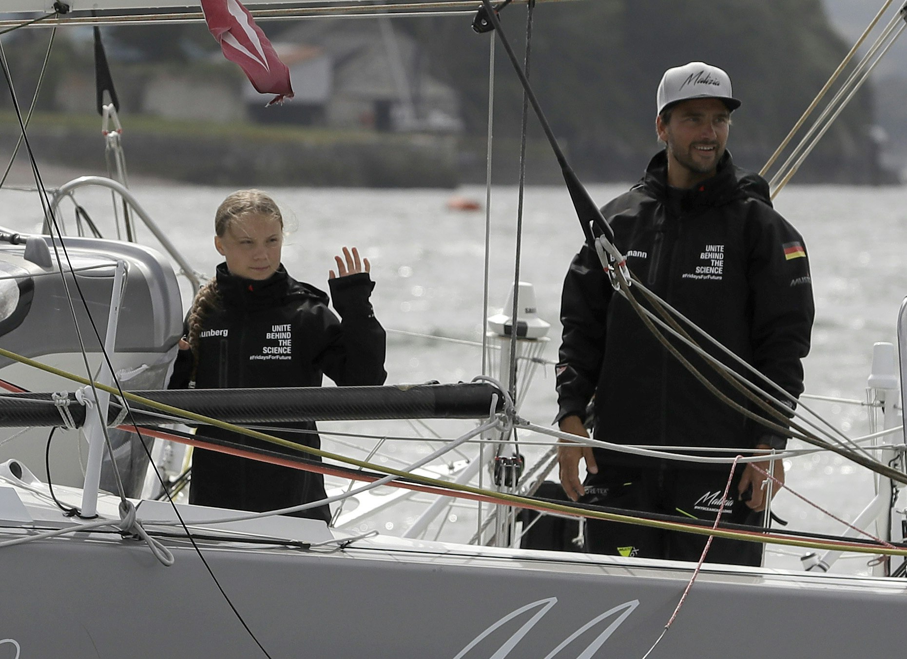 Climate activist Greta Thunberg stands on a solar powered sailboat ahead of her voyage to the 2019 United Nations Climate Action Summit. She wears a black windbreaker with the words United Behind the Science printed in white. She stands next to a man wearing a similar jacket, with the German flag on the left sleeve and a white ball cap on his head. 