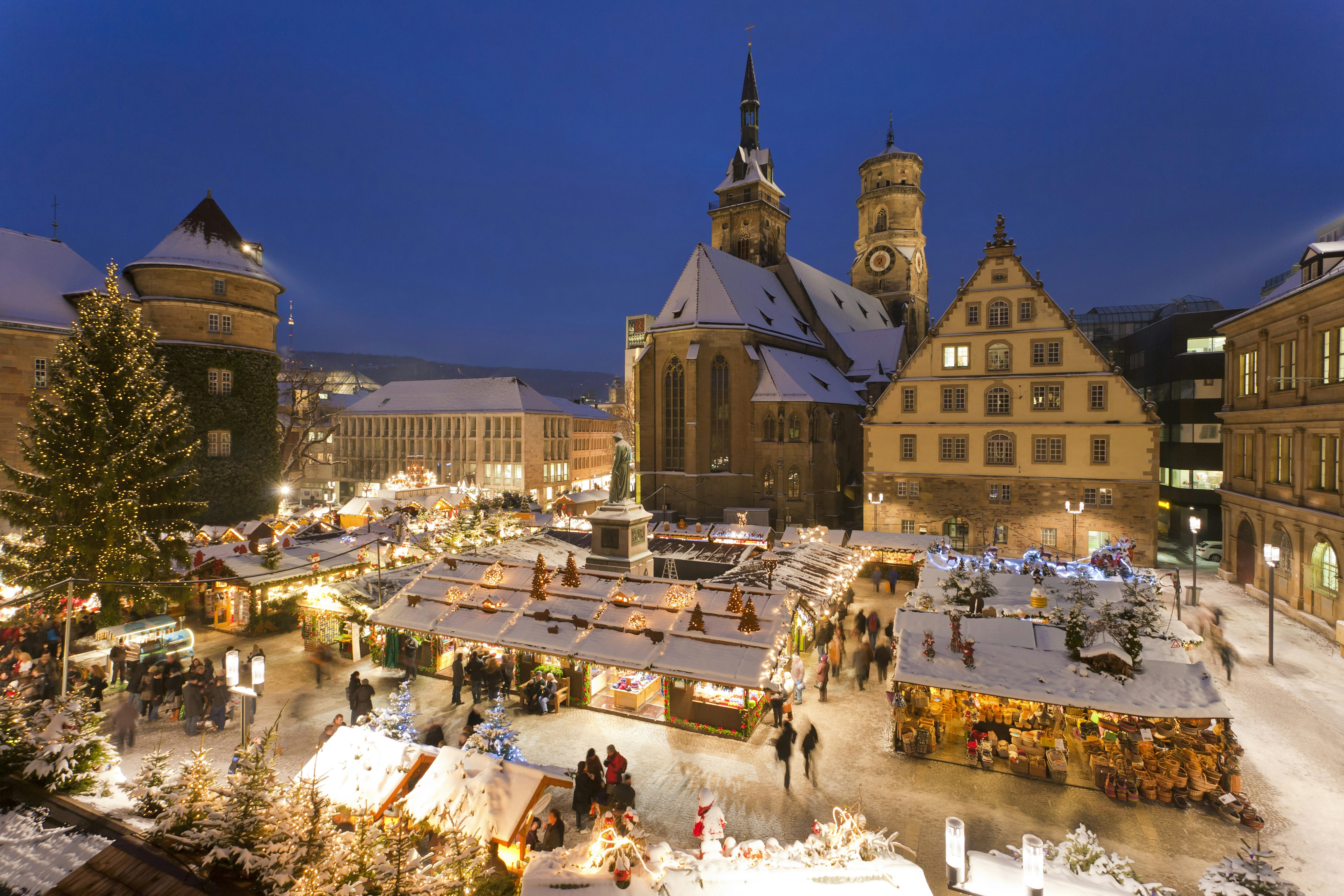 A snowy nighttime scene of a Christmas Market in a pretty medieval town square. A tree has been dressed with fairy lights and visitors walk between the many wooden stalls.