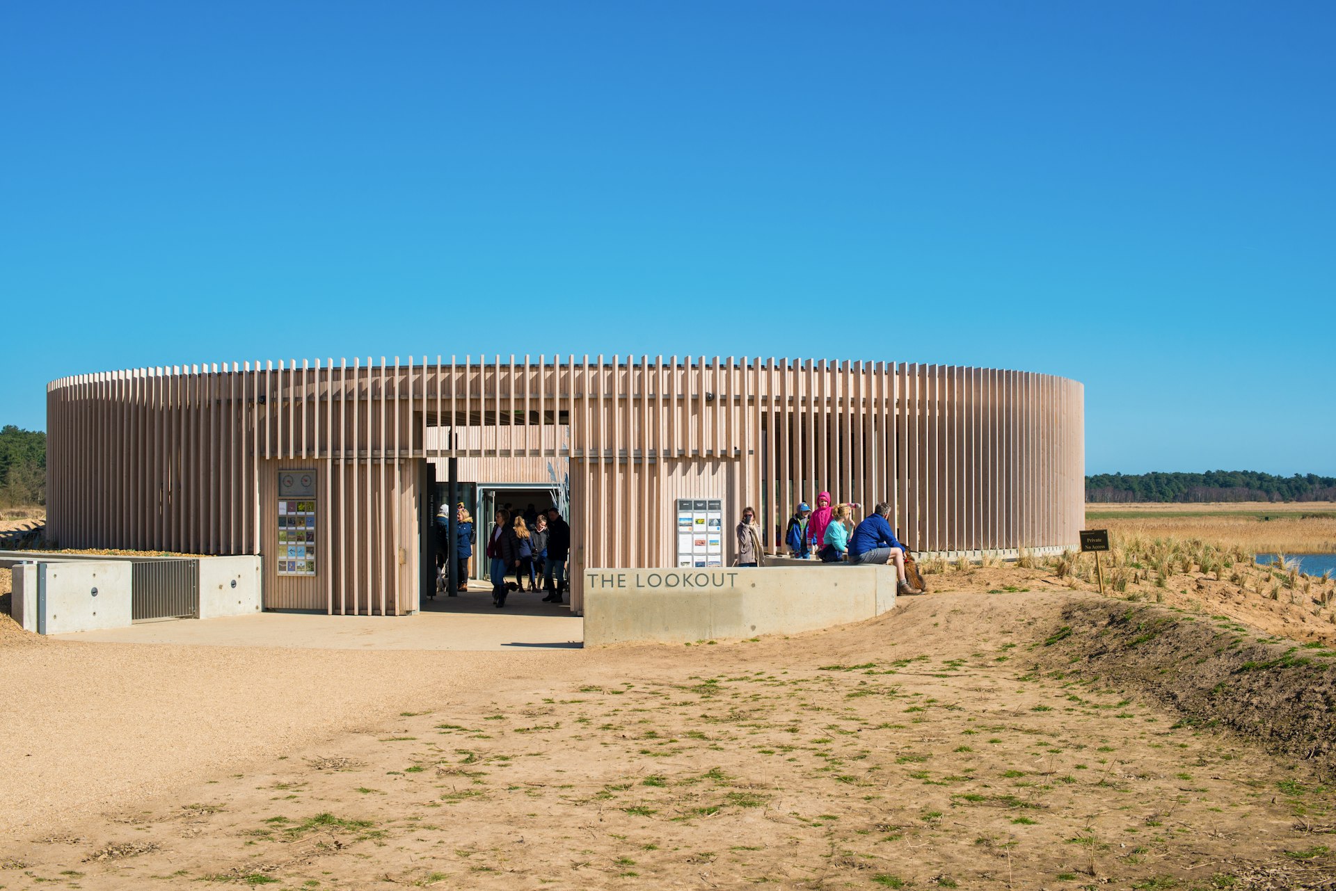 A circular, one-story building in a pinkish tan color with evenly spaced vertical strips of wood surrounding the exterior sits on a matching brown scrub plain in East Anglia, England. A row of tourists in colorful windbreakers sit on a low wall to the right, while just inside the door several figures stand in shadow. 