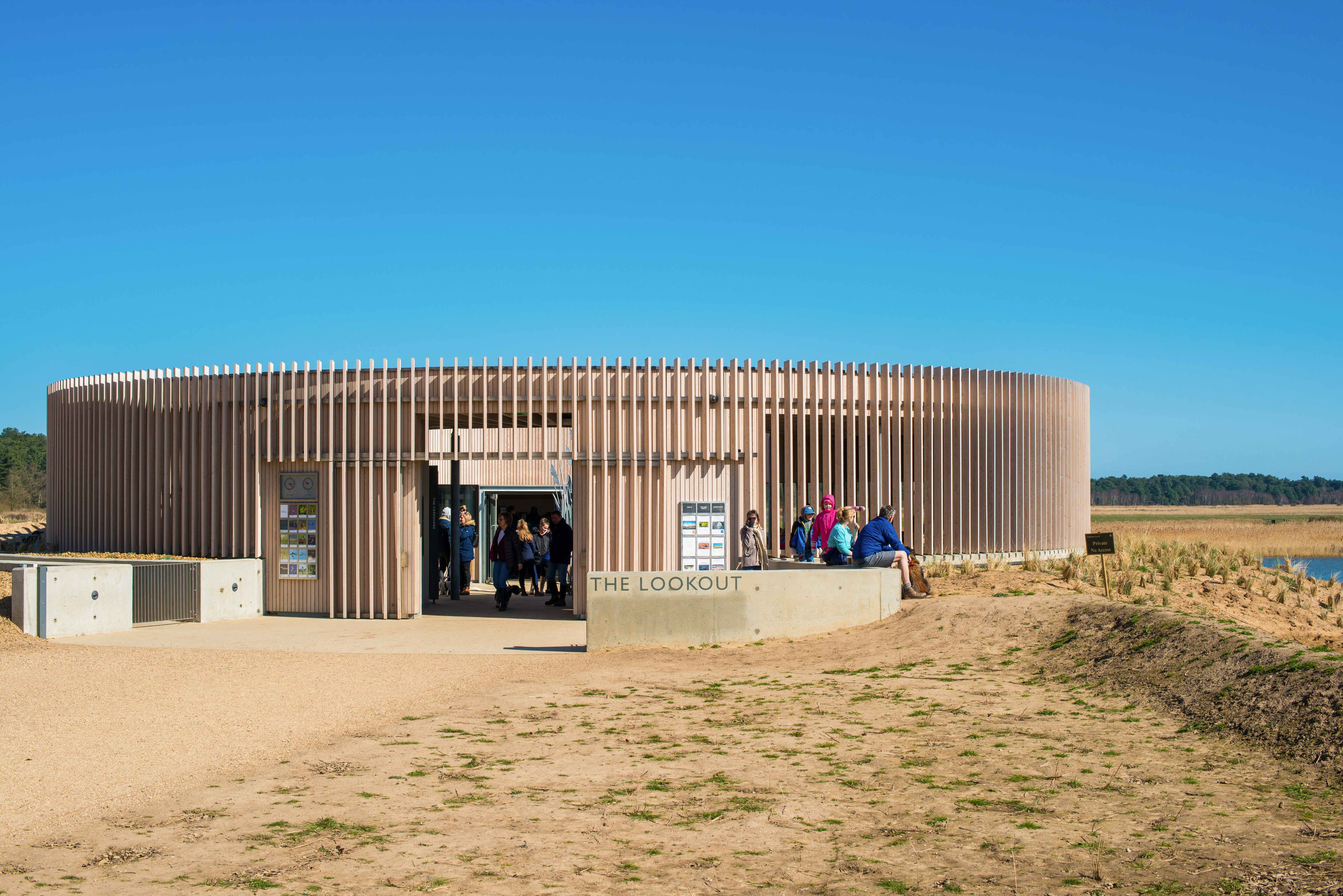 A circular, one-story building in a pinkish tan color with evenly spaced vertical strips of wood surrounding the exterior sits on a matching brown scrub plain in East Anglia, England. A row of tourists in colorful windbreakers sit on a low wall to the right, while just inside the door several figures stand in shadow. 